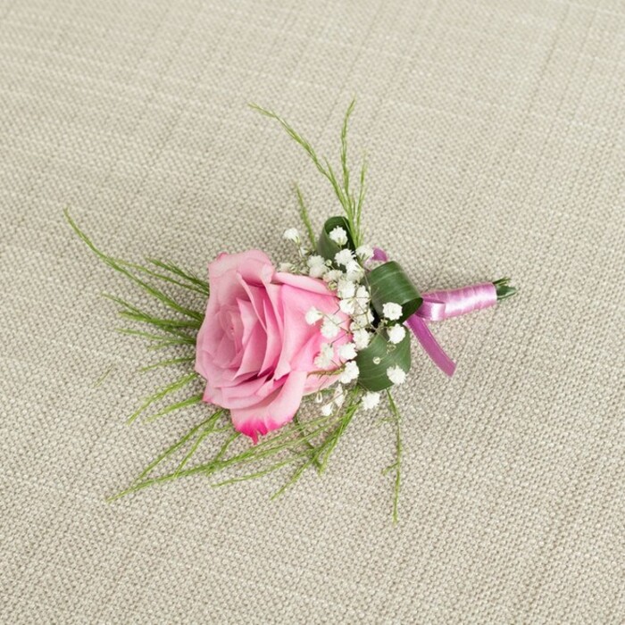 A meticulously crafted pink rose boutonniere rests on a light beige, naturally textured linen surface, the kind of calming neutral you might find in a Tufnell Park townhouse or a boutique venue off Brecknock Road. The image is captured from above, giving a clear, intimate view of every floral detail. The centrepiece is a single, exquisite pink rose, not fully open yet radiantly in bloom, with petals that spiral out from a softly blushed core to edges tinted with a richer, more saturated rose-pink. The velvety petals fold and curl gently, hinting at that just-cut freshness you'd expect from a local florist preparing flowers on the morning of a wedding. Encircling the base of the rose are airy tufts of white baby's breath, their tiny star-like flowers clustered so densely that they form a feather-light cushion around the main bloom, offering a delicate contrast in both texture and colour. Fine strands of narrow, grass-like greenery emerge from behind, fanning out in slender lines that give the boutonniere a sense of movement and grace. Tucked more closely to the stems, a broader, darker green leaf has been artfully folded, almost like a protective shell, lending shape and structure to the compact arrangement. At the base, the stems are wrapped in a lustrous satin ribbon, blending soft lavender-pink tones with deeper magenta-purple hints, secured in a simple yet precise knot that keeps the piece refined but not fussy. The gentle weave of the linen background is softly visible beneath, its understated pattern adding an organic, tactile context without distracting from the floral design. Light falls evenly across the composition, creating subtle highlights along the ribbon and delicate shadows between petals and foliage, giving this boutonniere the dimensional presence of a carefully considered finishing touch for a groom, groomsman, or prom guest heading out across Tufnell Park's tree-lined streets.