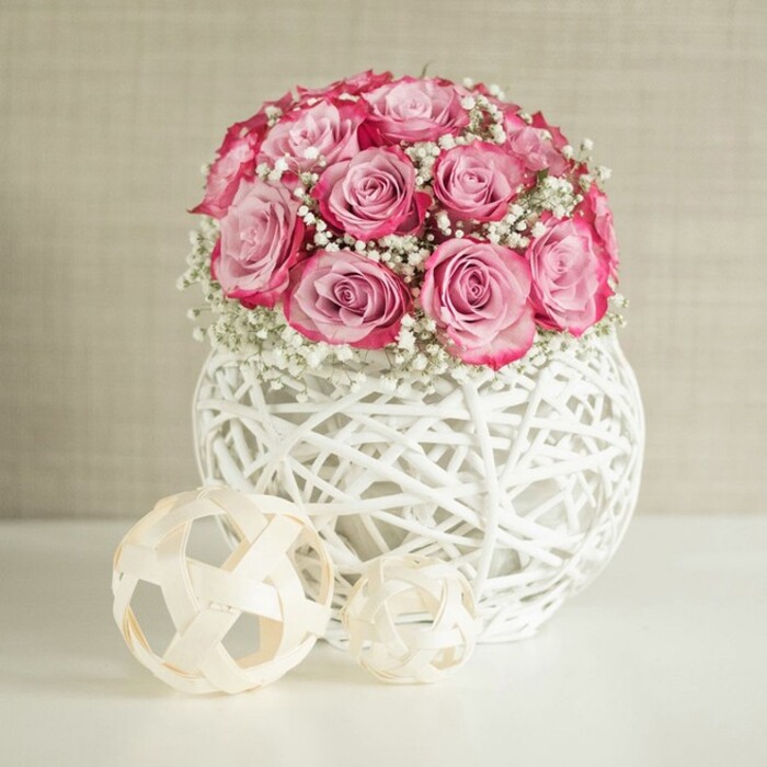 A carefully composed indoor scene presents a lush spherical bouquet of pink roses and white baby's breath in a modern woven vase, creating a look that would suit a stylish flat near Tufnell Park station. The bouquet itself is generously rounded, made up of around fifteen roses, each bloom fully opened to reveal layers of velvety petals. The colouring is nuanced: the centres are a soft, powdery blush, while the outer edges deepen into a richer, almost fuchsia pink, giving the arrangement a gentle ombré effect that draws the eye from the top of the dome down to its base. Among the roses, fine stems of gypsophila are deftly tucked, their clusters of tiny white blossoms adding lightness and volume, like delicate clouds floating between the more substantial rose heads. This mix of colour and texture evokes a sense of romance and freshness, perfect for anniversaries, new baby celebrations, or same-day delivery to homes and cafés throughout Tufnell Park. The flowers are gathered in a distinctive spherical vase fashioned from white-painted natural fibres, perhaps wicker or rattan, woven into an open, basket-like structure. Gaps in the weave let light filter through, creating intricate shadows on the pristine white surface beneath. To the left of the vase, two woven decorative spheres in a pale, natural wood tone sit on the tabletop, their open geometric forms echoing the texture of the vase while adding a cosy, Scandinavian-style touch often seen in local interiors around Lady Somerset Road. Behind the arrangement, a soft beige or off-white wall with subtle texturing provides a serene background, allowing the pink and white tones to stand out without distraction. The lighting is bright yet gentle, with no harsh contrasts, so the scene feels calm, polished, and quietly luxurious-like a moment of everyday beauty captured between busy North London days.