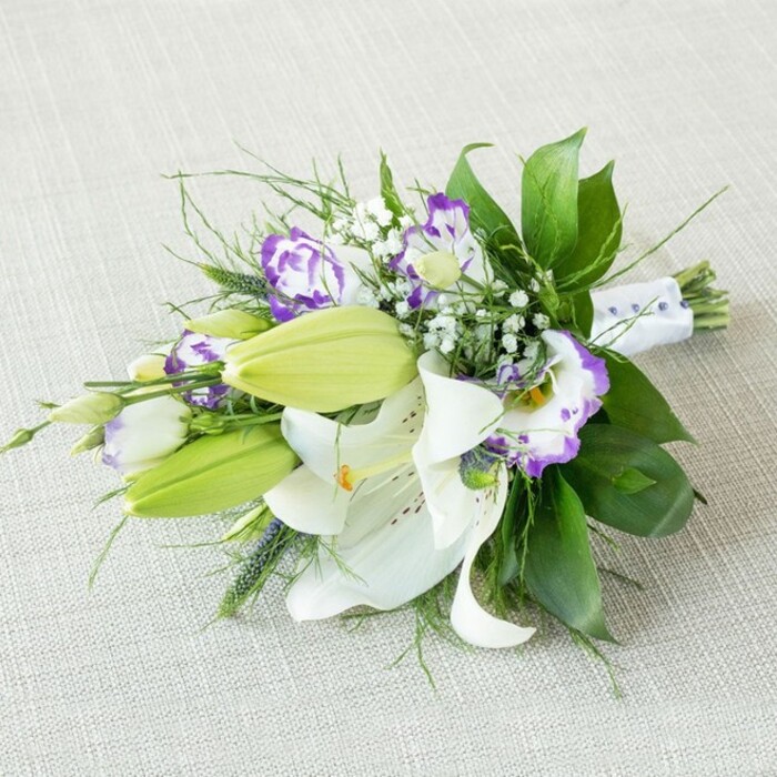 A refined bridal-style bouquet stretches diagonally across a softly lit, light beige fabric, its slightly nubby texture suggesting natural linen or cotton, creating a calm, understated stage for the flowers. Dominating the arrangement, several large Oriental lilies bloom in full splendour, their snowy white petals gently arcing backward as if opening to the light. Each lily reveals prominent yellow-orange stamens, dusted with pollen, and a delicate scattering of cinnamon-coloured speckles along the throat, lending the flowers a sense of depth and character. Alongside these open blossoms, elongated lily buds in a bright, spring green rest tightly closed, their smooth surfaces and pointed tips promising a longer life for the bouquet once it's in someone's hands in Tufnell Park or along nearby Fortess Road. Between the lilies, clusters of lisianthus unfurl like miniature, ruffled cups. Their petals are creamy white at the base, gradually deepening into rich purple edges that range from soft lilac to bold violet, creating a painterly effect that adds sophistication and contrast. Sprigs of gypsophila weave through the composition, their countless tiny white blooms forming cloud-like tufts, lightening the overall feel and creating a sense of air and space between the larger flowers. Surrounding all of this is a lush collar of foliage: broad, glossy emerald leaves provide structure and a deep green backdrop, while slender, more feathery greens and a few spiky accents extend outward, giving the bouquet energy and a natural, garden-inspired movement reminiscent of the planting beds you might see around local parks. At the narrower, right-hand end, the stems are gathered tidily and wrapped in a smooth white satin ribbon, which appears to be decorated with evenly spaced, pearl-like beads or small purple dots. This careful finishing touch speaks to a florist's practiced hand and suggests that the bouquet is designed for a special moment-perhaps carried down an aisle, placed as an elegant centerpiece at a celebration, or given as a thoughtful gift delivered to a doorstep in Tufnell Park, radiating freshness, grace, and quiet joy.