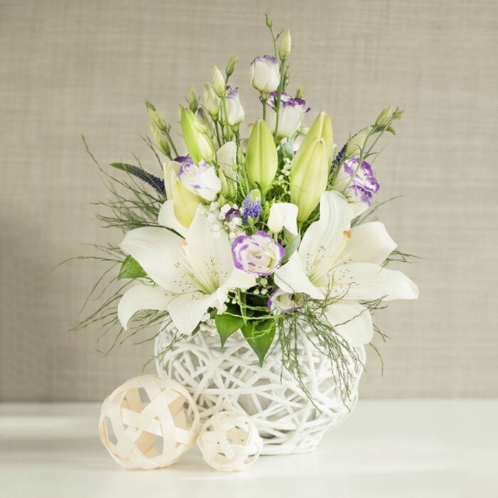 A refined floral arrangement in soft whites, greens, and touches of lavender stands poised on a crisp white surface, as if ready to be placed on a wedding reception table in Tufnell Park or along a quiet dining table near Lady Somerset Road. At its heart is a sculptural white vase, round and slightly squat, with an intricate open-weave pattern that resembles hand-woven rattan, letting the light pass through and create delicate shadows. From this vase, tall, graceful stems of white lilies emerge, some flowers fully open with wide, trumpet-like petals curving outward to reveal their central stamens dusted with warm pollen, others tightly enclosed in elongated light green buds that promise days of continuing bloom. Between the lilies, clusters of lisianthus offer a softer, ruffled texture: their mostly white petals are tinged with subtle lavender or pale purple along the edges or centres, creating a gentle watercolour effect. Some lisianthus blossoms are fully open, while others remain in bud, contributing to a sense of gentle unfolding. Scattered throughout are tiny white filler flowers, likely baby's breath, mingling with hints of deeper purple florets that add depth without breaking the calm palette. Lush green foliage, from feathery fern-like fronds to slightly broader, glossy leaves, spills and arches around the base, giving the design a natural, almost garden-like feel. In the foreground, two pale woven spheres in different sizes sit on the white surface, their open latticework echoing the vase's texture. A softly textured beige wall behind the arrangement provides a neutral, linen-like backdrop, enhancing the bouquet's tranquil, sophisticated character, ideal for serene celebrations, sympathy tributes, or quiet reflection at home in Tufnell Park.