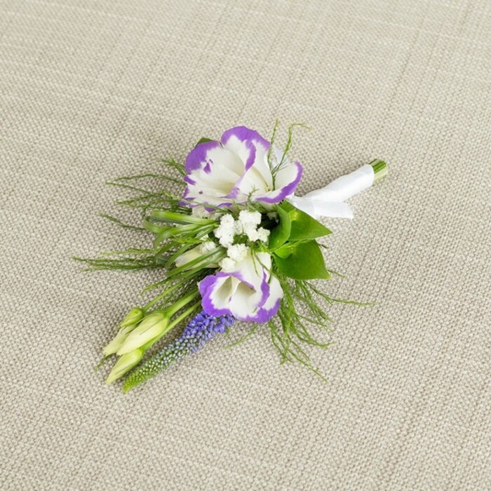 This image shows a thoughtfully designed boutonniere resting on a softly textured, light beige fabric background, likely linen, whose neutral colour allows every floral detail to stand out with clarity. The arrangement is centred around two striking lisianthus blooms, their petals a crisp, luminous white with richly ruffled edges brushed in a deep, regal purple, creating an elegant contrast reminiscent of the refined townhouses and characterful streets near Tufnell Park station. Surrounding these focal flowers are tiny clusters of pristine white filler blooms, similar to delicate baby's breath, which soften the composition and fill in gentle gaps with a misty, romantic haze. Rising gracefully among them is a slender stem of muscari, its closely packed, bell-shaped flowers shaded in an intense grape-purple, adding both height and texture to the buttonhole, like a vertical exclamation mark of colour. Fine, feathery greenery, likely asparagus fern, radiates around the flowers with a light, airy feel, while a few more solid green leaves offer structure and depth, evoking fresh foliage gathered from a well-kept garden off nearby Dalmeny Road. Two pale green buds remain attractively closed, hinting at the promise of blooms yet to open and subtly symbolising new beginnings. At the base, the florist has bound the stems meticulously with a smooth white satin ribbon, wrapped in even, careful turns and finished neatly to create a clean, polished handle for easy pinning. The atmosphere of the image is calm, romantic, and composed, suggesting a boutonniere suited to a groom or wedding party member attending a ceremony in Tufnell Park or nearby Gospel Oak, where understated luxury and thoughtful design are prized. It reflects the work of an experienced florist who understands how to balance colour, texture, and meaning in a small yet significant floral detail.