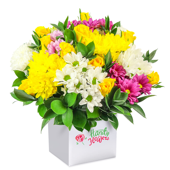A lush, dome-shaped floral arrangement overflows from a pristine white, cube-shaped gift box, photographed against a seamless white background so the intricate play of colours and textures is clearly visible. The bouquet is composed of a harmonious blend of yellow, white and fuchsia blooms, interlaced with deep green foliage to create a rich, garden-like effect that feels both modern and comforting, much like a bright corner café near Tufnell Park station. Dominating the design are large yellow chrysanthemums, their softly layered petals forming generous, cushiony blooms that radiate warmth. Nestled among them, small yellow spray roses and rosebuds provide texture and intimacy, some buds still tight and rounded, others just beginning to reveal their inner petals, suggesting early morning freshness. White flowers are thoughtfully scattered throughout: daisy-style chrysanthemums with vivid green centres lend a casual, meadowy charm, while fuller white chrysanthemums add a plush, cloudlike quality and increase the sense of abundance. Threaded between these are vivid fuchsia and deep magenta chrysanthemums, some with hints of lighter pink at the petal tips, injecting bold sparks of colour that echo the lively energy of nearby Holloway Road. The foliage, likely sturdy varieties such as ruscus, appears in glossy dark green sprays that peek between the blossoms and pool around the base, framing the entire display and providing a strong contrast to the lighter petals. The simple white box functions as a contemporary container and ready-made gift package, ideal for convenient delivery to homes and offices around Tufnell Park, Archway, and Gospel Oak. The overall mood is cheerful and uplifting, making this arrangement perfect for marking birthdays, saying thank you, or sending a burst of colour to brighten an otherwise ordinary day.