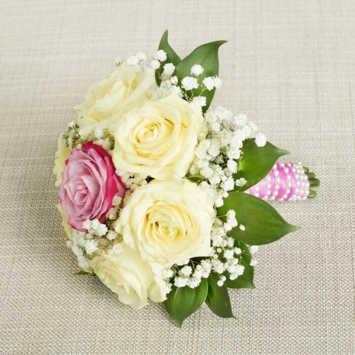 Viewed from above on a gentle angle, this sumptuously arranged bouquet radiates freshness and modern romance, its florals artfully presented against a soft, buff-toned linen backdrop. At its core lie five creamy roses, their petals opening with quiet anticipation-each one poised at the peak of bloom and reminiscent of the refined style so often admired on Fortess Road or in the intimate venues of Tufnell Park. Slightly offset amidst the pale roses, a vivid fuchsia-pink rose becomes the focal point, drawing in a viewer's gaze with its striking hue and symbolising passionate celebration. Threaded throughout, abundant clusters of white baby's breath disperse a light, feathery airiness that softens the stronger floral shapes and adds a timeless touch of grace. Lush, waxy green foliage anchors the bouquet, giving shape and depth, while the stems extend elegantly to the right, bound in brilliant fuchsia satin ribbon. This tactile ribbon, wrapped with care, is further adorned with a graceful coil of tiny iridescent pearls, adding a sophisticated shimmer that catches the shifting light. Resting on the textured linen-a surface evocative of artisanal textiles favored by Tufnell Park artisans-the bouquet stands as a testament to local floral design, perfect for weddings, anniversaries, or any event where classic elegance meets personal style. Every detail reflects the neighbourhood's creative spirit and the meticulous expertise of Tufnell Park florists.