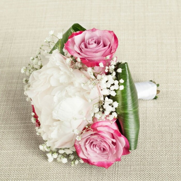 Captured in gentle, natural light that filters much like the ambience in hidden Tufnell Park courtyards, this compact bouquet exudes luxury and poise. Several flawless white peonies, their myriad ruffled petals reminiscent of clouds gathering before a spring sun, form a sumptuous base. Carefully interspersed are two radiant pink roses, their petals displaying a painterly transition from vivid fuchsia boundaries to soft blush-pink and whispery lavender hearts, each bloom catching the quiet magic of early mornings near Dartmouth Park Hill. The bouquet is sprinkled with ethereal gypsophila, its delicate white florets filling subtle gaps and infusing the piece with a hint of whimsy-much like the artistry found in Tufnell Park's galleries and gardens. The floral design is beautifully structured by a lush collar of folded tropical green leaves, such as ti or aspidistra, delivering bold definition and a sense of botanical richness. All is bound together with a seamless white satin ribbon, forming an inviting handle ready for a Tufnell Park bride or bridesmaid's gentle grasp. Resting upon textured, light beige linen evocative of local interiors, the arrangement's mix of textures-velvet petals, glossy leaves, frothy baby's breath-delivers a sophisticated yet natural elegance. Perfect for weddings, proms, or anniversary events across the neighbourhood, this bouquet speaks of heritage, artistry, and heartfelt celebrations crafted just steps from the Northern Line.