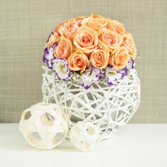 A graceful floral centerpiece is captured in soft, diffused light, as if it has just been placed on a prepared table for a celebration in Tufnell Park. The main focus is a lush, perfectly rounded dome of roses, each one a soothing shade of peach, apricot, or soft salmon. Their velvety petals spiral outward in tight, generous swirls, overlapping to form a smooth, continuous surface with no breaks in colour. This dense mass of roses feels opulent yet gentle, reminiscent of a summer sunset over leafy side streets near Tufnell Park Fields. Wrapped around the base of the rose dome is a halo of white lisianthus blooms. Their petals are finely ruffled and edged with rich violet and purple tones that look almost hand-inked, adding depth and a painterly contrast to the warm roses above. Nestled between the lisianthus are delicate sprays of gypsophila, their tiny white flowers scattering light like a fine mist, giving the entire arrangement a breathable, romantic quality. The bouquet is anchored in a distinctively shaped, broadly spherical container, crafted from white woven wicker or rattan. The open latticework design creates an airy, sculptural feel; glimpses of the interior shadows and stems add quiet intrigue and underline the artisan craftsmanship, much like the handmade pieces you might find in local boutiques around Fortess Road. In front of the main piece, resting on the same immaculate white surface, lie two decorative spheres fashioned from pale, natural wood-toned strips, interlaced to form open frameworks. The larger and smaller orb echo the texture and pattern of the vase, completing the composition with a touch of playful geometry. Set against a subtly textured beige background, the arrangement exudes calm sophistication and timeless romance, ideal for wedding table styling, anniversary dinners, or refined home décor in North London homes that appreciate soft colour and thoughtful detail.