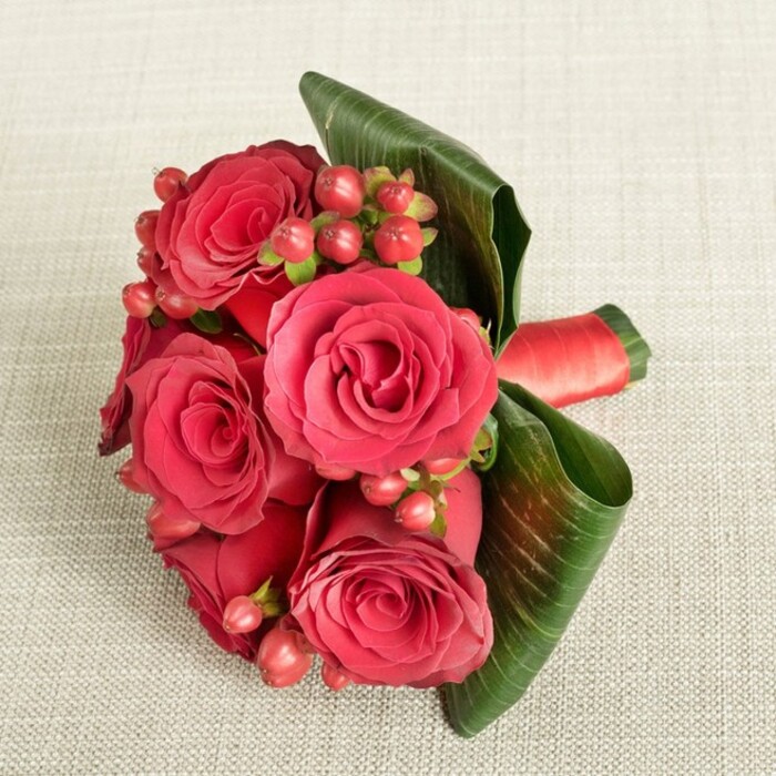 An elegant hand-tied bouquet of rich red roses and berries lies diagonally across a soft, light beige fabric, its finely woven texture providing a gentle, understated backdrop that lets the colours truly sing. The arrangement features around six full-headed red roses, each bloom beautifully open yet still structured, with inner petals forming a tight, velvety spiral that deepens to near burgundy at the centre, and outer petals relaxing outward into warmer scarlet tones. Between the roses, multiple clusters of small, rounded Hypericum berries in deep red and reddish-pink add a playful contrast, their smooth, glossy surfaces and tiny green stems bringing both texture and a slightly festive touch. Along one side, broad, dark green tropical leaves have been carefully curved and folded, hugging the bouquet like a sculpted frame, their subtle veins catching the light and adding depth. The stems are gathered neatly and secured by a wide, rich red satin ribbon, wrapped tightly to create a clean, tailored finish that continues the romantic colour scheme. The photograph is taken from a gently elevated angle, allowing you to see the full face of the roses and the intricate mingling of blooms, berries, and foliage while the neutral fabric beneath softens any shadows. The lighting is even and flattering, highlighting the natural sheen of the leaves and the plush, almost touchable texture of the petals. The overall impression is one of intimate sophistication, perfect for an anniversary, a proposal, or a special date night in Tufnell Park, perhaps after an evening wander past the quiet garden squares and leafy residential streets that characterise this North London pocket.