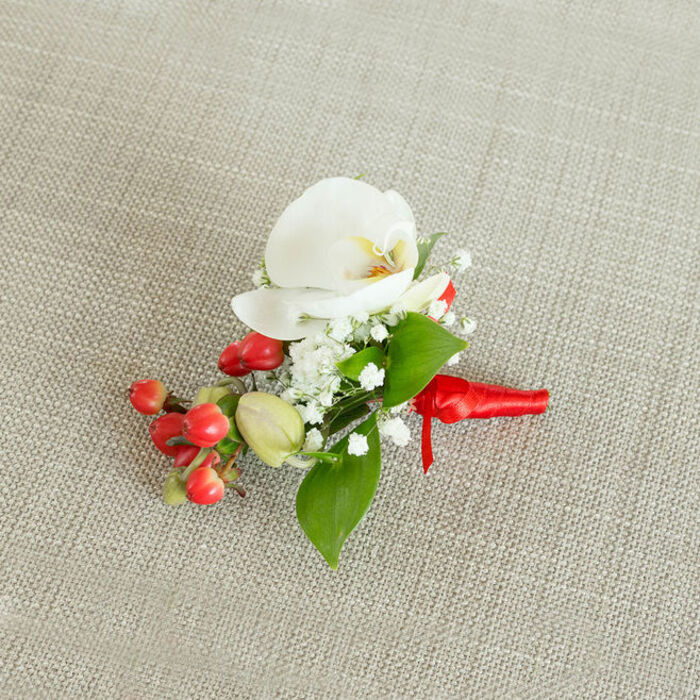 A refined floral boutonniere is captured from above, resting on a light beige, linen-like fabric whose subtle texture recalls the calm, understated charm of Tufnell Park terraces off Brecknock Road. The eye is immediately drawn to the central bloom: a pristine white Phalaenopsis orchid, its wide, velvety petals unfolding in a gentle arc. Within the open flower, a hint of buttery cream and soft orange glows at the throat, adding warmth to the otherwise pure white palette. Gathering around the orchid, countless tiny white blossoms of baby's breath weave a delicate halo, their fine stems and cloud-like clusters giving the composition an ethereal softness. To one side, a striking group of glossy, round red berries sits like drops of lacquered colour, bold yet elegant, suggesting winter celebrations and candlelit receptions in nearby Islington and Kentish Town. A few smaller, pale green buds nestle alongside, lending a sense of freshness and subtle movement. Green foliage of varied shapes-some broader and spear-like, others slim and elongated-threads through the flowers and berries, providing structure, contrast, and a natural frame that makes the white orchid stand out even more. The stems are neatly gathered and wrapped in a rich red satin ribbon, its smooth surface catching the light. The ribbon is tied into a small, secure knot, with short, tidy tails that rest neatly against the woven fabric backdrop, hinting that the piece is ready to be pinned to a lapel or worn as a delicate corsage. The overall mood is one of quiet sophistication and heartfelt celebration, a floral detail that would feel perfectly at home at a wedding or formal event in and around Tufnell Park, where thoughtful, handcrafted touches are treasured.