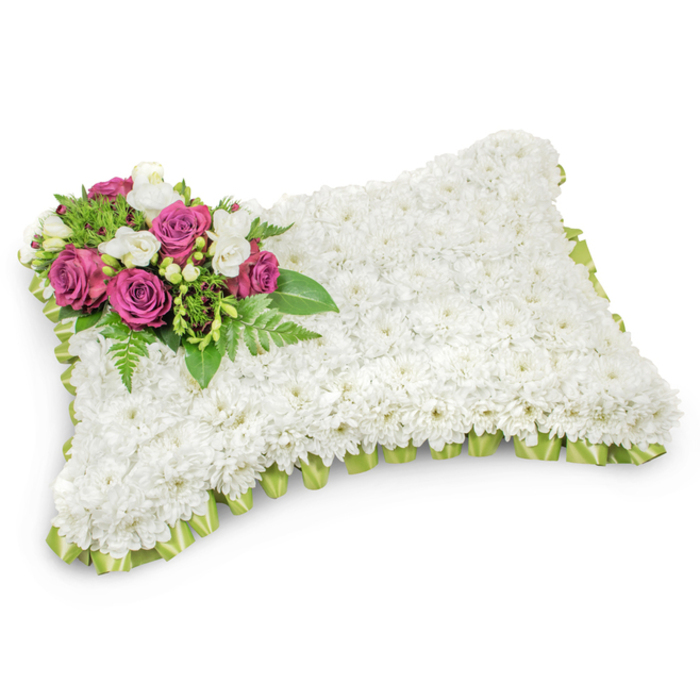 A traditional rectangular funeral pillow, rendered with meticulous floral technique and set against a neutral white backdrop to emphasise form and detail. The principal plane of the tribute is a continuous field of immaculate white chrysanthemums, each bloom densely clustered so the surface resembles a plush fabric pillow-soft to the eye and suggestive of peaceful repose. Crowning the upper-left corner is a composed spray of deep magenta and fuchsia roses, their velvety spirals providing a poignant accent of colour and reverence. Sprigs of small white freesias or like blooms nestle between the roses, their delicate shapes and faint fragrance bringing a quiet brightness and innocence to the cluster. Supporting foliage-feathery fern fronds and glossy green leaves-fans beneath the accent, offering contrast and the feeling of a living foundation. The perimeter is neatly finished with a pleated, light green satin ribbon that gently emerges beneath the white carpet of blooms, lending a subtle outline and a hint of pastoral green. Under soft, diffused light the textures translate as tactile: the chrysanthemums feel like a linen cushion, the roses like velvet, and the ribbon like a gentle fold. This arrangement reads as a composed expression of sympathy suitable for a chapel or graveside service in and around Tufnell Park and nearby Dartmouth Park; the piece is designed to communicate peace, respect, and lasting remembrance in a form both familiar and refined.
