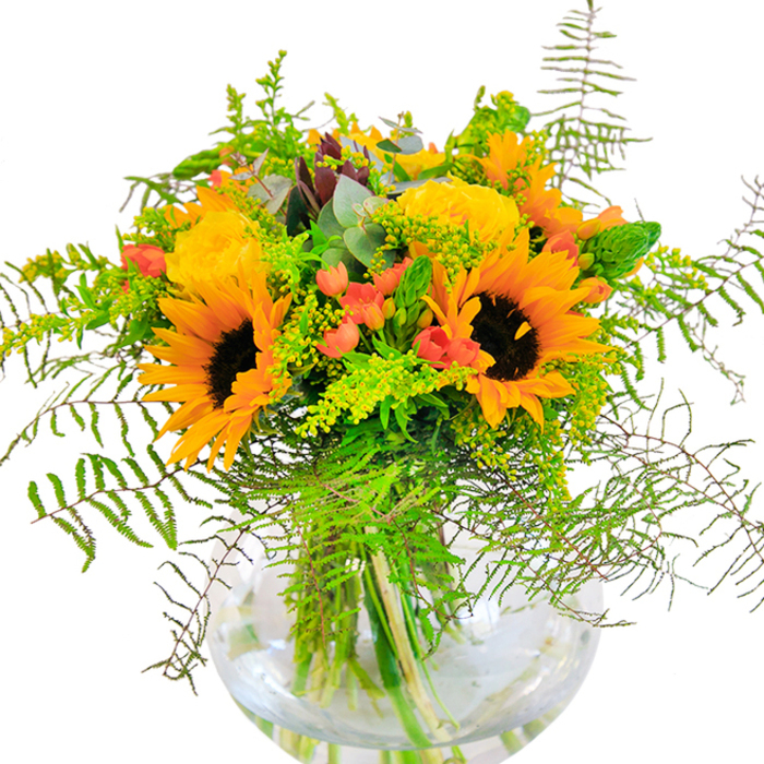 Bathed in the soft, natural light that recalls early afternoons in Tufnell Park, this abundant bouquet is a radiant expression of happiness and comfort. Sunflowers stand proudly at the heart, their golden petals wide and bright, echoing the warmth that fills the local cafes and parks. Tucked between their sunlit faces, lavish yellow roses spiral open into velvety blooms, their subtle fragrance mingling with the fresh scent of cut greenery. Pops of vivid red hypericum berries catch the eye, nestling among gentle sprays of feathery ferns and light-green filler that softens the whole composition with playful movement. Slender green stems reach upward alongside silver-blue eucalyptus, offering textural contrast and a hint of urban wildness-reminiscent of Highgate's woodland edges. All elements are intimately gathered in a round, clear glass vase, where the stems form graceful lines beneath the surface, illuminated by the water's reflective glow. This joyful bouquet, perfect for celebrating birthdays, anniversaries, or welcoming new neighbours along Tufnell Park Road, embodies the energy and friendliness of the neighbourhood. It's a floral gesture that brings the outdoors in, filling any room with light, warmth, and a sense of long summer days in North London.