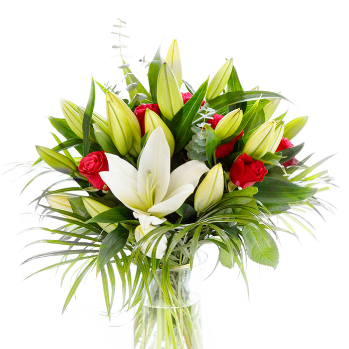 This photographed bouquet is anchored by a single, prominent white Oriental lily in full bloom, its creamy, slightly translucent petals opening to reveal pale yellow stamens and a light green pistil at center stage. Encircling the focal bloom are numerous lily buds in lime-green to pale yellow tones-some tightly closed, others beginning to unfurl-creating layered rhythm and the suggestion of gradual revelation. Nestled between these are small, velvety red roses and rosebuds, their deep crimson hues mostly restrained within curled petals, delivering traditional romance and visual contrast without overwhelming the arrangement's serenity. The foliage selection amplifies texture: broad, dark forest-green leaves provide structure and shadow; long, delicate palm-like fronds in emerald green cascade and soften the bouquet's silhouette; and slender stems of silvery-green eucalyptus with rounded leaves lend a muted, cool breathing space among the denser greens. The flowers are composed in a clean, cylindrical clear glass vase, with pale green stems visible below the waterline-an intentional transparency that speaks to freshness and skilled handling. Shot against a brilliant white background, every contour, shadow, and subtle colour shift is clarified, making this ideal for product imagery or as a visual reference for clients in Tufnell Park seeking an elegant gift or centerpiece. The overall impression is one of tasteful celebration and refined sympathy at once: suitable for anniversaries, weddings, or quiet remembrances, where the purity of the white lilies and the passion of the red roses combine to communicate deep feeling with grace.