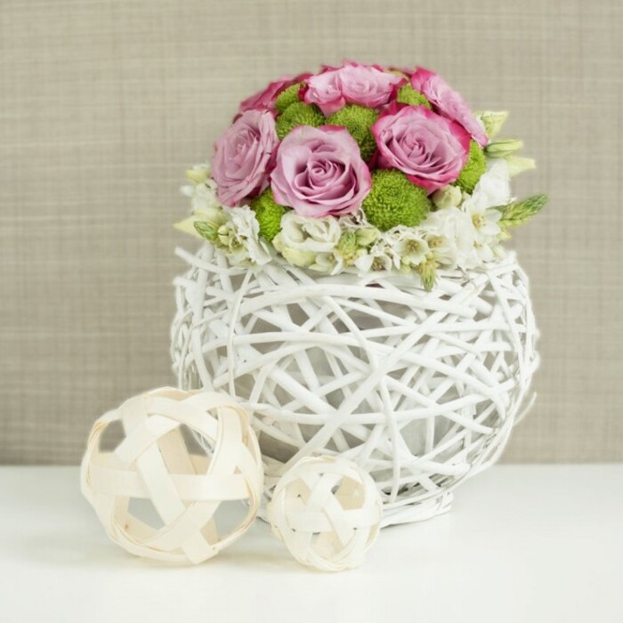 A refined floral composition sits serenely on a crisp white tabletop, the kind of arrangement that could anchor a wedding table setting in Tufnell Park or a celebratory brunch near Holloway Road with understated sophistication. At the centre of the scene is a spherical white vase, cleverly constructed from thin, branch-like strips woven into an open wicker design, creating the look of a modern, airy basket with gentle gaps that invite light and shadow to play across its surface. From this sculptural base rises a compact, rounded bouquet overflowing with fresh roses in shades of pale pink and soft lavender, their velvety petals forming intricate spirals and rosettes. Many of the blossoms display deeper pink edges, adding subtle depth and a painterly quality to the overall colour palette. Nestled among the roses are clusters of bright green button mums, their tiny, rounded blooms offering a vivid, grassy contrast that enlivens the arrangement and draws the eye inward. Around the perimeter of the floral dome, delicate white filler flowers-small, starry blooms that could be waxflower or similar-peek through the larger heads, softening transitions and lending a cloud-like lightness to the design. To the lower left of the vase rest two decorative spheres of woven natural wood or rattan, one slightly larger than the other, their pale, straw-like strands looping and interlacing to create open, sculptural forms that echo the vase's texture. Behind it all, a softly blurred background in muted beige tones, reminiscent of linen or lightly textured fabric, provides a calm, neutral canvas. Gentle, diffused lighting highlights each petal and twig, conveying a sense of freshness, craftsmanship, and quiet celebration, as if prepared by a seasoned local florist for same-day delivery to homes and venues across Tufnell Park and nearby Gospel Oak.
