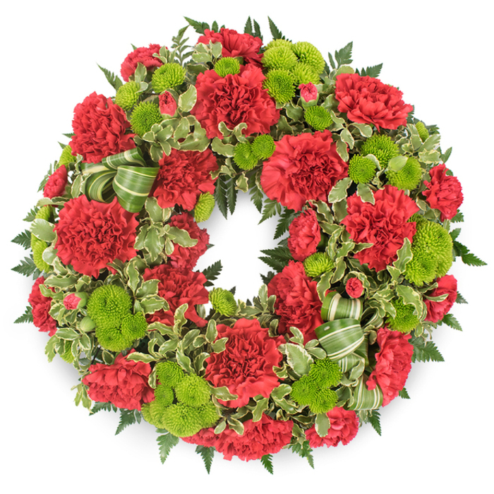A vividly coloured circular wreath, designed as a heartfelt funeral or memorial tribute, is set against a clean white background so that its rich reds and luminous greens seem to float in the air, much like a striking arrangement you might see delivered to a service in Tufnell Park. The wreath's structure is heavily packed with deep red carnations, arranged with precision yet allowed to retain their natural, slightly irregular forms. Each carnation is built from layers of finely frilled petals, giving the surface of the wreath a soft, almost velour-like texture. The reds range from warm crimson to slightly deeper, wine-toned shades depending on how the light catches them, evoking a sense of gravity and enduring affection. Punctuating this sea of red are clusters of lime green button chrysanthemums, also known as pompon mums. These tiny globes of petals are tightly composed, each one almost perfectly spherical, standing out like fresh green beads between the larger carnation heads. Their bright chartreuse colour introduces a vibrant, uplifting contrast that keeps the wreath from feeling sombre, suggesting renewal and hope. Encircling and weaving among the flowers is a mix of lush greenery: dark, feathery fern fronds anchor the design with depth and shadow, while variegated pittosporum, its leaves edged in soft cream, lends a lighter, patterned accent reminiscent of the varied foliage found in local parks such as Tufnell Park Playing Fields. A decorative ribbon in shades of green, with diagonal striping, is thoughtfully tucked into the lower area of the wreath, forming a few elegant loops that echo the circular form without overshadowing the blooms. This carefully crafted display, created with the practiced eye of a neighbourhood florist, would sit beautifully beside a coffin in a Tufnell Park church or at a crematorium nearby, speaking quietly of remembrance, gratitude, and a life cherished.