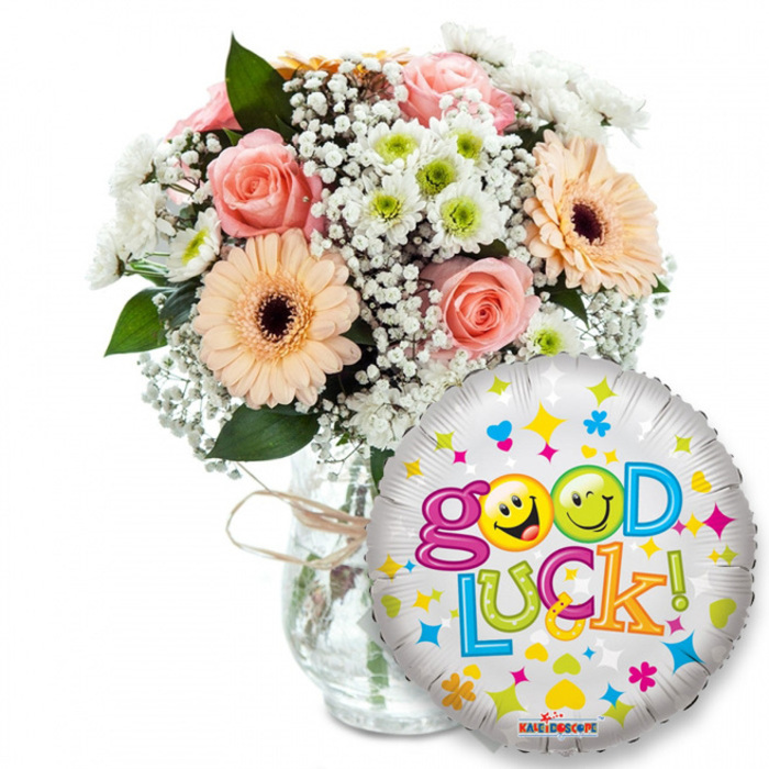 A tender, upbeat 'Good Luck!' duo arranged with florist care and local sensibility from Flowers Tufnell Park, pictured against a stark white backdrop so every colour and texture is clear. The bouquet, positioned slightly left of centre, is assembled in a clear glass vase whose visible water and stems speak to freshness; a rustic twine looped around the vase neck completes the handmade feel. Peachy-orange Gerbera daisies with their dark brown centres bring sunny faces to the design, while pale pink roses, their petals tightly coiled, supply a soft romantic note and a gentle fragrance. Billows of white Gypsophila (baby's breath) lace through the blooms, creating airy movement, and clusters of small white and lime-green chrysanthemum florets add crisp highlights and a hint of verdant sparkle. Rich green foliage peeks from beneath, grounding the colourways and adding depth under soft studio lighting. To the right and slightly in front of the vase floats a round metallic silver Mylar balloon, patterned with multicoloured stars, hearts and confetti-like shapes, and emblazoned with the buoyant message 'Good Luck!' in a bubbly, rainbow sequence of letters. The two 'o' letters in 'Good' are cheerful yellow smiley faces - one winking, the other sticking its tongue out - which gives the gift a playful, personal touch; a small KALEIDOSCOPE logo is visible at the balloon's base. This pairing reads as the perfect local send-off for someone heading to university, starting a job near Upper Holloway, or taking any new step, and it reflects the calm, expert hand of a neighbourhood florist.