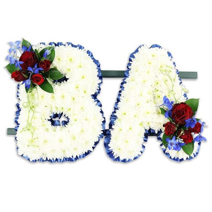 A carefully composed floral tribute spells the initials B A in bold capital letters, each letter formed from a tightly packed mosaic of tiny pristine white chrysanthemums whose layered petals create a soft, clouded texture and a feeling of serene purity. The edges of both letters are accentuated with a luxurious royal blue satin ribbon, its ruffled banding giving definition and a respectful formality to the shapes. The B carries a lush accent at its upper left curve-a cluster of deep red roses in mixed stages of bloom paired with bright, spiky blue delphinium-like flowers and framed by dark green foliage and pale green trailing stems-while the A is balanced by a mirrored cluster at its lower right. Those red and blue accents are arranged to catch the eye without overpowering the white base, their colours reading as love and steadfastness set against calm white. The pair rests together on a slim, dark green rectangular base, displayed side-by-side as if two names standing in quiet solidarity. The neutral white background keeps the focus entirely on the craftsmanship: the fine textural play of chrysanthemums and the velvet of roses, the sheen of ribbon, the cool pop of blue spikes. As a florist with experience delivering memorial pieces to local venues around Tufnell Park, Junction Road and the surrounding Camden area, I recognise the balance of composition and symbolism here-an arrangement designed to convey respect, memory and a very particular personal connection through carefully chosen colour and form.