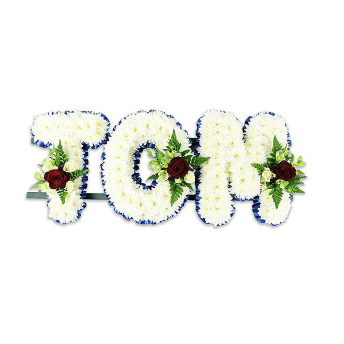 A dignified, personalised memorial piece that spells TOM in bold, three-dimensional letters, each completely covered in neat clusters of pristine white chrysanthemums. As a Tufnell Park florist familiar with local services around Tufnell Park Road and Dartmouth Park, I picture this arrangement standing at a chapel near the tube or at a local crematorium: the chrysanthemum faces are packed closely to create a plush, voluminous texture that reads both soft and substantial. The edges of every letter are traced in royal blue ribbon, a refined outline that sharpens each curve and adds a calm, respectful contrast to the pale blooms. Each letter bears a small, symmetrical spray featuring a single deep red rose as its visual anchor, accompanied by smaller ivory spray roses and feathery fern fronds; these accents introduce warm colour and gentle foliage movement against the white. A slim dark metal support is visible beneath the letters, providing stability while remaining unobtrusive. Photographed on a spotless white background with diffused daylight, the piece suggests stillness and careful craft-the scent of roses faint in the air, the texture of chrysanthemum petals soft beneath the eye. Prepared with attention to detail and sensitivity to family wishes, this bespoke name tribute is suited to funerals or memorial services across Tufnell Park, Archway and nearby Camden, and can be arranged for delivery locally by Flowers Tufnell Park.