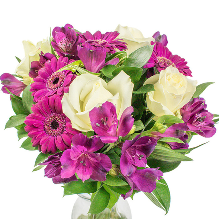 Framed against a pure white studio background, this compact, round bouquet reads as a study in colour harmony and florist craftsmanship, the kind we prepare at Flowers Tufnell Park for local clients picking up near Tufnell Park station, Dartmouth Park Road, or the nearby market. The eye is immediately drawn to several large magenta Gerbera daisies whose vivid, fuchsia-leaning petals fan out from dark, textured centres, projecting a lively, contemporary energy. Interwoven with these are creamy white roses - their petals layered and soft, releasing a muted, classic scent that tempers the gerberas' brightness - and occasional Lisianthus-style ruffled blooms that bring a subtle bell-shaped elegance to the midplane. Clusters of deep purple Alstroemeria, the Peruvian lilies, provide complexity: trumpet-like petals with delicate speckling and darker streaks that invite a closer look and lend the bouquet a hint of exotic refinement. Lush, dark green foliage - broad leaves alongside narrower, sword-like greens - forms a verdant frame that sculpts the bouquet into a full, rounded silhouette and creates pockets of shadow and light. A faint edge of a clear glass vase at the base anchors the composition, its suggestion of water reinforcing the freshness of each stem. Bathed in soft diffuse lighting, the colours pop crisply against the white background. This arrangement feels perfectly suited for birthdays, thank-you gifts, or a thoughtful housewarming present in Tufnell Park, and demonstrates the tactile, sensory approach our local floristry team uses when choosing colour, scent, and seasonal texture.