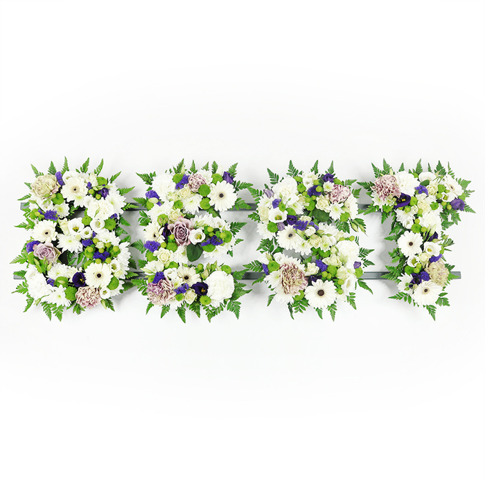 An elegant, horizontal set of four floral letters spells BEST, each character arranged with meticulous care against a bright white surface. The composition leans heavily on serene whites - prominent, pristine Gerbera daisies with their darker central discs anchor the design alongside creamy lisianthus and full, peony-like white carnations that lend a soft, layered texture. Interspersed are soft mauve and lavender roses and ruffled carnations that introduce touches of warmth and solemnity; tiny deeper-purple filler blossoms nestle into shadowed pockets to add definition and visual depth. Along every contour, feathery green fern fronds form a natural, scalloped border, their airy plumes softening the letter shapes while small green button chrysanthemums dot the interior as lively, fresh punctuation. A dark structural base provides subtle contrast where it shows through, giving each letter a firm silhouette without stealing attention from the flowers themselves. The overall effect is quietly reverent - the whites glow with a gentle luminescence in soft daylight, while the purples read like respectful accents. This is the kind of personalised floral lettering often chosen for memorial services, chapel tributes, or cemetery placements around Tufnell Park and nearby Dartmouth Park or Holloway Road, where friends and family gather to honour a life. The craftsmanship is visible in the balance of textures and in the way scent and colour work together to form a composed, heartfelt message of love, respect, and enduring remembrance.