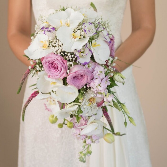 In this evocative bridal bouquet scene, a bride from Tufnell Park stands with graceful poise, showcasing a lavish, waterfall-style arrangement that feels plucked straight from a Hampstead Heath meadow on a spring morning. Her arms encircle the bouquet with a gentle reverence, drawing the eye to the dramatic cascade of pristine Phalaenopsis orchids, their broad white petals kissed with golden centres, arranged to spill luxuriously downward. Surrounding the orchids, full-bloom blush pink and lavender roses offer soft, romantic hues, mingling with the subtle notes of powdery fragrance often found in North London's local flower markets. Wisps of delicate baby's breath float among the larger blooms, gifting the composition an ethereal, almost otherworldly edge, while sprigs of lavender and purple blossoms-perhaps stock or sweet pea-add shading and nuance to the colour story. Trailing fuchsia amaranthus strands sweep gracefully, their textured magenta length resembling wild tresses in the breeze, bringing an artistic sense of drama and movement. Polished green foliage and the occasional bud emerging from the bouquet's core evoke freshness and craftsmanship, as if arranged only moments ago for a ceremony in Tufnell Park's historic churches. The creamy, soft-focus background ensures the bride's finely patterned lace gown and the floral masterpiece are visually elevated. Every blossom in this bouquet narrates a tale of love, creativity, and wholehearted celebration, embodying the refined taste of Tufnell Park's vibrant community.