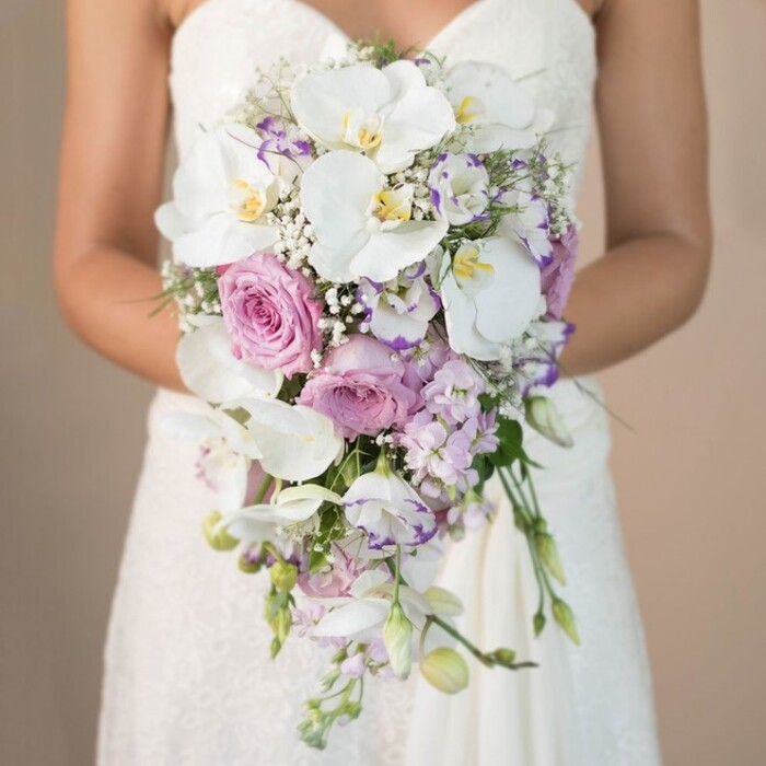 This richly detailed, close-up image showcases a bride-only visible from the chest down-standing in the heart of Tufnell Park and holding a luxurious cascading bouquet against a smooth, neutral backdrop. Her classic strapless wedding dress, fashioned in intricate ivory lace, hints at timeless elegance echoed in historic local venues. The central highlight of the bouquet is a series of immaculate white Phalaenopsis orchids, their generous petals open wide, each bloom revealing golden centers and a whisper of lavender veining. Enveloping them, plush pink and blush garden roses nestle tightly, their velvety spirals reminiscent of fresh spring mornings in Tufnell Park's community gardens. Scattered throughout are gentle lavender stock and delicate lisianthus-some ruffled and edged in deepest purple-providing a whimsical counterpoint. Baby's breath, threaded with care, fills spaces with ethereal softness. A touch of trailing greenery and unopened buds complete the bouquet's organic, flowing silhouette, drawing the eye downward in a cascade of lush, harmonious colour. The clean, subdued background sets a restful, sophisticated tone, allowing each floral element to shimmer. This bouquet, created for local couples seeking the finest wedding florals in and around Fortess Road or the lively borders of Kentish Town, is a testament to classic romance and artistic floral craft. Every bloom, every twist of lace, and every whisper of scent captures the storybook magic unique to Tufnell Park celebrations.