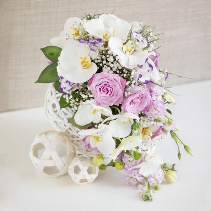 Centred on a spotless white tabletop, a lavish floral arrangement captures the eye with its soft, romantic palette and sculptural textures, reminiscent of a carefully styled wedding table in a Tufnell Park townhouse. Dominating the bouquet are several impressive white Phalaenopsis orchids, their arching stems and broad, silken petals creating a graceful, winglike form. Each blossom's centre glows with a subtle mix of lemon-yellow and warm gold, adding depth and an air of quiet luxury. Cradling these orchids are plush roses in pastel pink and pale lavender, some fully open to reveal intricate, swirling layers of petals, others just beginning to unfurl, suggesting freshness and movement. Threaded throughout, smaller ruffled flowers with white and purple or fuchsia-edged petals, likely Lisianthus or Eustoma, provide an artistic, dappled effect, much like glimpses of colour in Waterlow Park in early summer. Wispy sprays of white baby's breath are scattered generously, their tiny, cloud-like blooms giving the arrangement a dreamy, airy lightness. From the lower edge, slender stems of delicate, light purple blossoms cascade down, softening the outline and introducing a gentle waterfall effect that would suit an intimate Tufnell Park wedding reception. Lush green foliage, in various shapes and sizes, peeks through the flowers, adding structure and a fresh, botanical scent to the imagined scene. All the stems gather into a striking spherical vase composed of interwoven white strips, its open, basket-like shell resembling an oversized ball of fine wicker. Placed next to it on the white surface are two matching decorative orbs in a lighter cream tone, one larger and one smaller, echoing the vase's texture. A softly textured, neutral beige background keeps the focus firmly on the flowers, enhancing the sense of calm sophistication, celebration, and refined local craftsmanship.