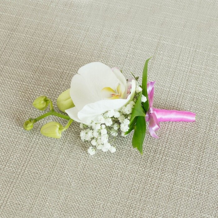 This image shows an exquisitely crafted boutonniere arranged horizontally on a softly textured, light beige linen-like fabric, the neutral background chosen to emphasise the calm, refined beauty of the flowers. Dominating the composition is a single white Phalaenopsis orchid, its broad, velvety petals spreading out in a smooth, symmetrical curve that feels both modern and timeless. The orchid's centre reveals delicate details: a wash of soft yellow in the throat, with the slightest flush of pale pink, giving the bloom an almost glowing heart when touched by the gentle, even lighting. Nestled closely around the orchid's base are airy clusters of white baby's breath, each tiny flower forming part of a cloudlike mass that brings texture, volume, and a sense of quiet romance reminiscent of intimate gatherings in Tufnell Park's townhouses and local venues. Slender, vibrant green leaves slip out from behind the floral focal point, their fresh colour sharply contrasting with the whites and creams and echoing the greenery of nearby streets like Carleton Road. Off to the side, a small branching stem holds a couple of tightly closed lime-green orchid buds, their smooth, rounded shapes signalling promise and future blooming moments-an understated metaphor for new beginnings and celebrations. The stems are expertly bound together with a bright fuchsia pink satin ribbon, tied into a small, perfectly formed bow whose lustrous sheen adds a striking, joyful accent, ideal for a groom's lapel, a groomsman, or a stylish prom attendee. Crafted with the care you'd expect from a local Tufnell Park florist, the boutonniere feels intimate, elegant, and quietly celebratory, ready to accompany a special outfit with graceful understatement.