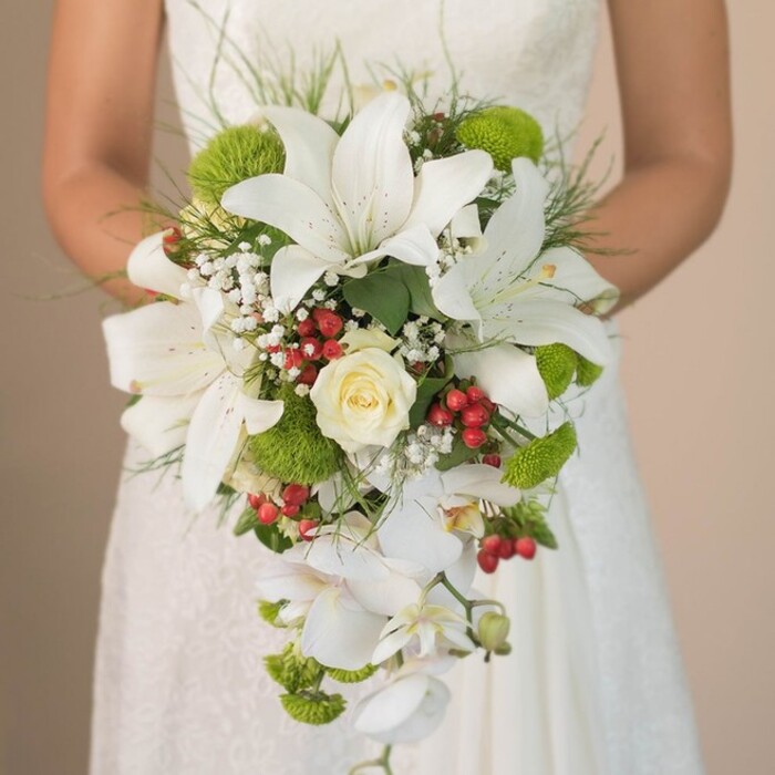 A close, chest-level portrait captures a bride holding her cascading bouquet just below the bodice of her delicate lace gown, against a softly blurred background that hints at Tufnell Park's tranquil charm. The bouquet's lush composition draws the eye with its majestic white lilies, their petals flecked with subtle maroon, radiating understated elegance. A central creamy rose, classic and finely furled, infuses the bouquet with time-honoured romance, while scattered sprays of white baby's breath introduce airy, cloudlike softness. Intertwined are clusters of lime-green cushion chrysanthemums and pom-pom flowers, their textured spheres bringing a refreshing, contemporary pop reminiscent of the vibrant greenery in Tufnell Park's community gardens. Vibrant red Hypericum berries dot the arrangement, offering a touch of festivity and colour reminiscent of autumn markets along Tufnell Park Road. Cascading below, a flourish of white Phalaenopsis orchids creates a striking waterfall, lending grace and movement to the silhouette. Wispy, grass-like foliage and trailing green leaves frame the bouquet, evoking the lush wildness of the local parks. The bride's gentle hold, hands dusted with sunlight, sets a mood of quiet anticipation, perfectly captured in the filtered daylight that highlights every floral nuance. This exquisite combination of classic and modern, expertly arranged with attention to detail by talented florists serving Tufnell Park, embodies a modern North London wedding-ideal inspiration for couples seeking luxurious, nature-infused bridal styles or exploring the latest in opulent floral trends.
