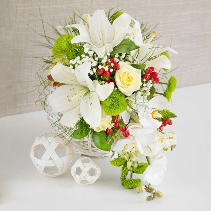 A beautifully composed white and green table arrangement, ideal for a modern Tufnell Park wedding reception or elegant dinner, sits proudly on a gleaming white surface against a softly textured beige backdrop. The flowers are arranged in a low, round white wicker basket whose subtle weave adds a natural, tactile foundation to the refined design. Dominating the upper part of the composition are several large white lilies, their trumpet-shaped petals fully open and gracefully curved, displaying faint freckles and long stamens topped with pale anthers, conveying both purity and drama. Just off centre, a single creamy ivory rose is nestled among the lilies, its soft yellow undertone providing gentle warmth and visual balance. From the right side of the arrangement, a flowing trail of white phalaenopsis orchids descends in a delicate arc, each orchid bloom pristine and smooth-edged, creating a classic cascading effect often chosen by brides in the Tufnell Park and Kentish Town area. Around and beneath these focal blooms, rounded bright green pom-pom chrysanthemums and tufts of fine, feathery greenery create a lush underlayer, while longer blades of ornamental grass add lift and movement. Tiny sprays of white baby's breath thread through the design, giving it a light, cloud-like softness, and clusters of deep red berries punctuate the whites and greens with small bursts of colour reminiscent of holly on winter strolls along nearby Dartmouth Park. To the left of the basket, two decorative white spheres made from interwoven strips of material rest side by side, one slightly larger than the other, echoing the basket's texture and lending an artful, sculptural touch. The whole scene is lit with gentle, even light that enhances the fresh, just-arranged look of the flowers, capturing a mood of quiet celebration and refined romance suited to intimate gatherings across Tufnell Park.