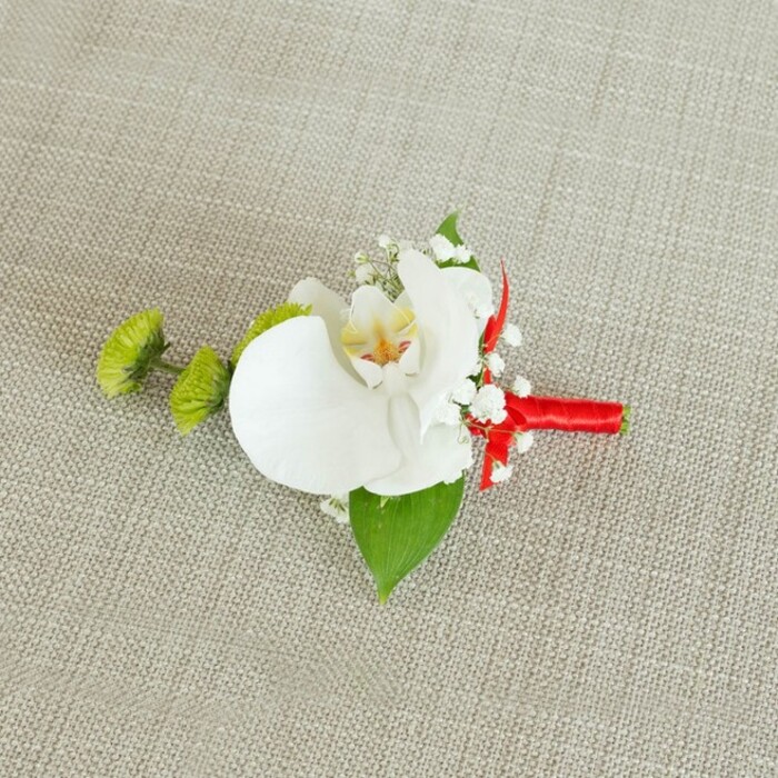 Displayed against a softly textured, light beige linen background, this refined boutonniere captures the quiet elegance of a Tufnell Park wedding morning. The fabric beneath has a slightly coarse, natural weave, like a favourite linen napkin laid out on a wooden table, providing a calm, neutral stage for the flowers to shine. At the centre rests a magnificent white phalaenopsis orchid, its petals broad and gently curved, their surface smooth and almost creamy in appearance. The flower's throat draws attention inward with delicate tones of soft yellow that fade into fine reddish-orange or pale brown markings, adding depth and subtle warmth to the bloom. Surrounding the orchid are airy tufts of white baby's breath, each tiny blossom contributing to a cloud-like halo, giving the piece an ethereal, romantic softness. On the left side, two small spherical lime-green chrysanthemum buds stand out with their intricate, slightly spiky texture, offering a contemporary flash of colour and echoing the fresh greens you might see along nearby Dartmouth Park Hill in early spring. A rich, deep green leaf forms a protective base beneath the main flowers, while another smaller, darker leaf close to the stems adds layers of greenery and structure. The stems themselves are bound with a striking crimson red ribbon, wrapped smoothly and firmly, then finished in a petite, decorative bow or knot that introduces a bold note of celebration. The lighting is gentle and even, letting the contrasts between white, green, and red remain crisp yet soothing. As a whole, the boutonniere feels both classic and lightly modern, a thoughtful floral detail perfectly suited to a groom, best man, or guest arriving at a local North London ceremony, lovingly echoing the understated style of the area.