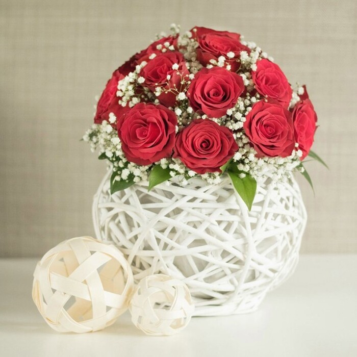 A carefully styled wedding table arrangement features a generous bouquet of deep red roses, displayed with understated elegance that would feel perfectly at home in a Tufnell Park townhouse. The roses are arranged in a rounded, almost spherical shape, blooms gathered closely together so that their velvety petals create one continuous surface of rich crimson. Each flower appears at its peak, with spiralled centres and gently curling outer petals that hint at a soft, classic rose fragrance. Threaded through this sea of red are fine sprays of tiny white baby's breath, their miniature blossoms adding both lightness and texture, like flecks of snow drifting across a velvet cloth. Subtle touches of green foliage are tucked between the blooms, visible but never dominant, reinforcing the sense of natural freshness. Supporting the flowers is a distinctive white vase, rounded and slightly bulbous, crafted from pale strands that weave and twist around each other in an open, basket-like pattern. This lattice structure gives the vase a handcrafted, contemporary feel, reminiscent of artisanal pieces you might find in small design shops off Junction Road. In the foreground, two decorative spheres made from similar woven material in soft beige tones rest on a smooth, pale tabletop. One is larger and placed slightly ahead of the other, creating depth and a gentle rhythm to the scene. The background is a simple, softly textured wall in light cream or sand, blurred enough that nothing distracts from the floral focal point. Soft, even light falls over the arrangement, enhancing the interplay of red, white, and neutral tones. The overall effect is one of quiet romance and refined celebration, perfectly suited to wedding receptions, anniversary dinners, or elegant gatherings in and around Tufnell Park.