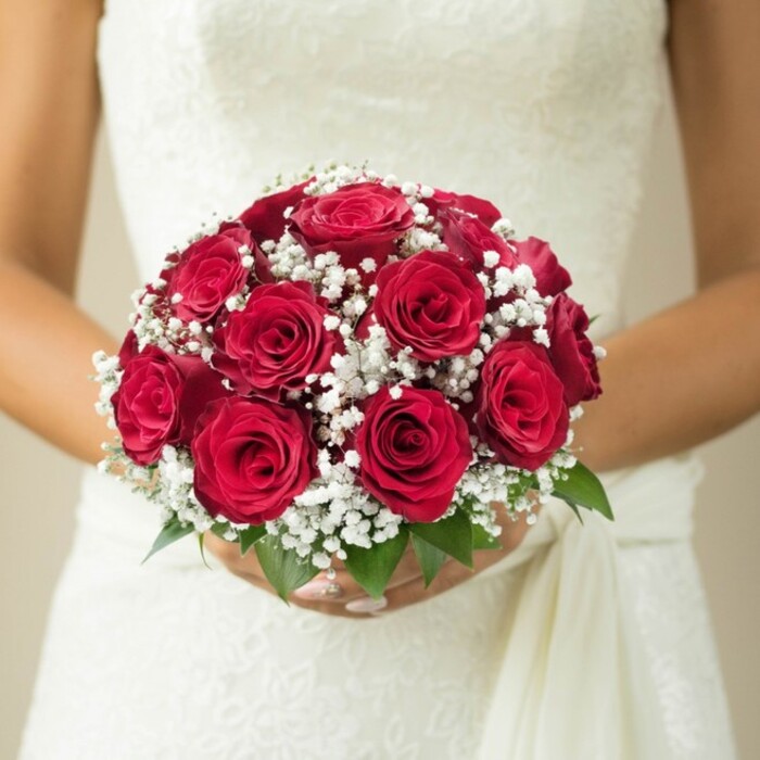 Capturing the spirit of a Tufnell Park wedding, this evocative image shows a bride-visible from the chest up-tenderly holding a stunning, traditional bouquet reverberating with the deep reds of classic roses. Each rose is artfully arranged, their tight, velvety petals forming a sumptuous, rounded cluster that speaks of love's enduring promise. Woven through the roses are generous clouds of white gypsophila, their delicate blossoms offering a feather-light softness and an airy touch that lifts the arrangement, a sight reminiscent of delicate wildflowers glimpsed near the paths of Tufnell Park itself. At the edges, bright green leaves break through, their natural freshness providing a subtle yet vital accent. The bride's graceful, manicured fingers envelop the stems, her pristine wedding gown-a tapestry of textured, potentially lace-embellished fabric-serving as a luminous canvas for the vibrant blooms. A draped sash or flowing swath of material descends from her right, adding movement and elegance to the composition. The background is muted-a soft gradation of cream and beige-evoking the gentle light of a Tufnell Park terrace and allowing the intricate details of the bouquet and bridal attire to truly shine. This bouquet is a timeless symbol of romance and tradition, perfect for wedding celebrations at local venues like the Star of Kings, and a testament to the craftsmanship local florists bring to every special day in Tufnell Park.