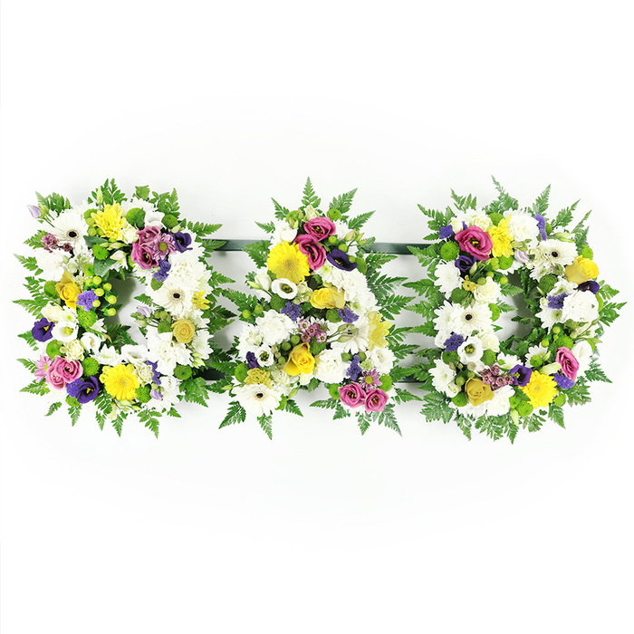 A heartfelt floral tribute spelling DAD is shown in crisp detail against a stark white studio background, the simplicity of the setting making every colour and texture more pronounced. Three individual letter frames, each one free-standing, form bold capital shapes lined with deep green fern fronds that curl outward in soft, natural arcs. The interior of each letter-the first 'D', the central 'A', and the final 'D'-is densely packed with fresh, front-facing blooms. At the base of the design are clean white chrysanthemums and broad white gerbera daisies, their layered petals overlapping to create a smooth, continuous surface that reads clearly from a distance. Among the whites are bright yellow gerbera blooms, petite yellow spray roses, and tiny button-like chrysanthemums that bring a gentle, hopeful warmth. Threaded through this lighter palette are rich purple lisianthus and small clusters of magenta or deep pink spray roses, plus hints of lavender-toned flowers that add depth and a more contemplative note, much like the quiet atmosphere in Tufnell Park's residential streets after a funeral service. Small mounds of fresh green button chrysanthemums echo the surrounding foliage, tying the colours together. Narrow, dark support bars link the letters discreetly along the back, giving strength while keeping the illusion of three separate floral blocks lined up side by side. The overall mood is dignified and deeply personal, a classic sympathy tribute a family might choose for a father's funeral or memorial gathering in Tufnell Park, expressing love, respect, and cherished memories in a single, clear floral word.