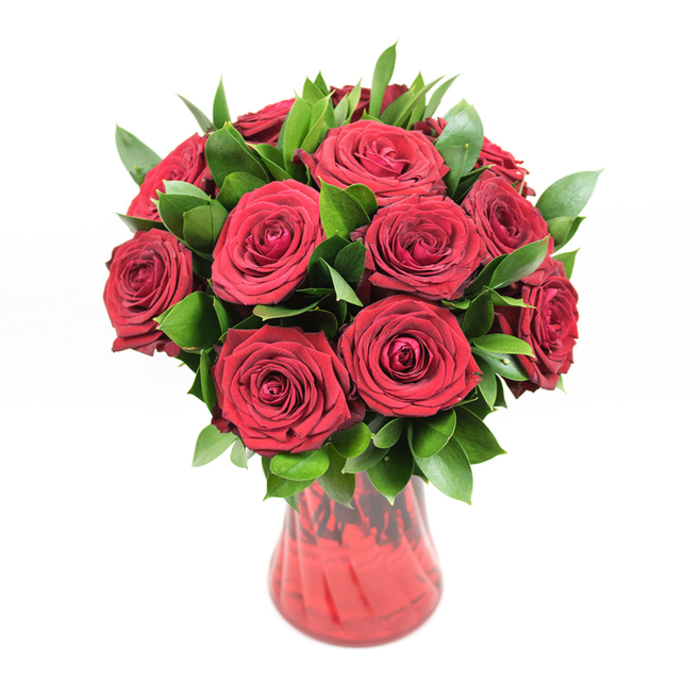 A classic round bouquet of about a dozen deep crimson roses is showcased in a translucent red glass vase, set against a bright white studio backdrop that highlights every curve and colour. The roses form a full, dome-like cluster, each bloom carefully positioned so their velvety petals open outwards in precise spirals, filling the arrangement with rich texture and depth. Their hue is an intense, romantic red, the sort that immediately suggests passion and lasting affection. Around and between the roses, ample dark green foliage-most likely ruscus or a similar leaf-adds fullness and definition. The leaves are smooth and slightly waxy, with a deep, glossy sheen that catches the light and frames the scarlet petals, preventing the arrangement from feeling flat. The bouquet appears expertly balanced, with no gaps or awkward angles, just a harmonious blend of flower and greenery. The vase itself is a key part of the composition: a red glass vessel with a gently flaring neck and a curving body that narrows towards the base, featuring a faint ribbed or swirled texture. This texture catches highlights and shadows, making the glass appear layered and alive, while its transparency allows you to glimpse the stems gathered neatly inside. With the neutral white background and even lighting, there are no distractions-only the interplay of red roses, green leaves, and red glass drawing focus. The overall feel is luxurious yet timeless, the kind of bouquet you might see on a polished table in a Tufnell Park townhouse near Tufnell Park Road, ready for an anniversary dinner, a romantic proposal, or a special same-day delivery to someone cherished. It conveys love with a quiet, confident intensity.