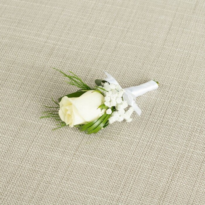 A meticulously crafted boutonniere is displayed on a gentle diagonal across a softly textured, light beige surface, the fabric's subtle linen-like weave adding a natural, almost homespun warmth. At the centre of the design sits a single creamy white rosebud, its petals closely wrapped in smooth, velvety layers that hint at a forthcoming bloom. A touch of soft green at the base of the rose-where the sepals cradle the bud-emphasises the flower's freshness, conjuring the feeling of a rose cut that morning, perhaps from a delivery arriving early to a florist just off Campdale Road in Tufnell Park. Surrounding the rose, airy clusters of white baby's breath extend in delicate sprays, each tiny blossom forming part of a gauzy, cloud-like collar that softens the overall outline of the boutonniere. Intermingled among these blooms, several sprigs of greenery emerge: some are slender and deep green, with neat, pointed tips, while others are more feathery and textured, reminiscent of fern fronds. Together, they add depth, gentle movement, and an organic contrast to the glowing white petals. The stems are gathered tightly and wrapped in a band of pure white satin ribbon that has a subtle sheen, reflecting the soft, diffused lighting in the scene. Just below the floral head, the ribbon is finished with a tidy bow whose symmetrical loops and short, crisp tails demonstrate careful, practised hands, like the signature finishing touch from the team at Flowers Tufnell Park. The lighting is even and flattering, with minimal shadows, allowing every fold of ribbon, every petal edge, and each tiny baby's breath bloom to be seen clearly. The boutonniere exudes traditional wedding elegance and would sit beautifully against a dark suit lapel for a ceremony at a local registry office or an intimate celebration in a Tufnell Park townhouse.