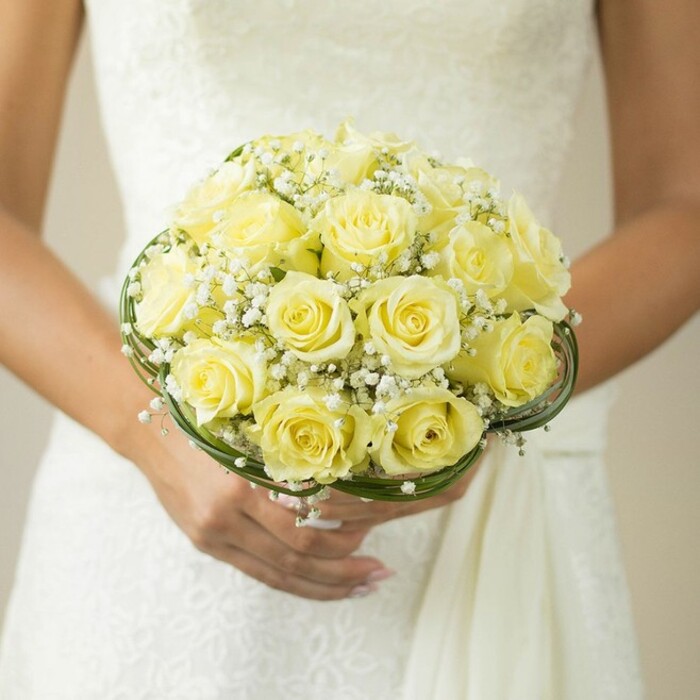 A close, tender view captures a quintessential Tufnell Park bridal bouquet, its gracefulness accentuated by the natural textures often found at local garden parties or Islington celebrations. The bouquet, perfectly rounded and densely arranged, is made up of numerous pale yellow roses, each bloom a symbol of happiness and new beginnings, their velvety petals opening in gentle spirals. Between the roses linger clouds of tiny, pristine white baby's breath, adding a softer dimension and a timeless elegance reminiscent of English spring mornings in Dartmouth Park. At the base, slender whorls of dark green leaf and delicate decorative wirings gently encircle the composition, giving it both structure and sophisticated charm-a technique favoured by experienced florists in the Tufnell Park community. The bride's hands, resting against the subtle white texture of a classic gown-perhaps detailed with lace or careful embroidery-cradle the flowers with reverence, her left ring finger adorned in simple promise. The background is thoughtfully blurred, ensuring attention is lavished upon the bouquet's glowing roses and the interplay of soft white and rich foliage. This evocative portrayal exemplifies the romance, craftsmanship, and meaningful elegance brought to wedding celebrations in Tufnell Park, perfect for those seeking inspiration for their own floral journey through north London's vibrant heart.