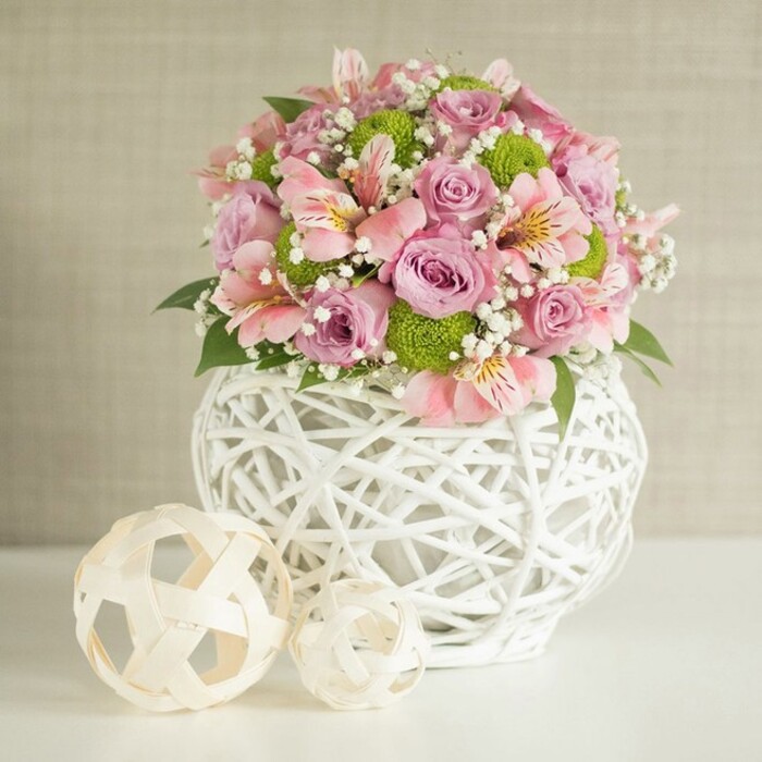 Brimming with soft elegance and local character, this floral centerpiece evokes a sense of welcoming luxury, reminiscent of Tufnell Park's mix of community warmth and modern style. Gracefully arranged on a flawless white table, the rounded bouquet bursts with pale pink and lavender roses, their plush, layered petals complemented by the delicate, trumpet-shaped blooms of pink alstroemeria featuring signature golden and fuchsia stripes. Pops of vivid lime green from compact button chrysanthemums, affectionately called 'Kermit' mums, add playful contrast and a modern twist. Interwoven sprigs of baby's breath, with their tiny white blooms, create a light, ethereal veil, while dark, glossy foliage grounds the entire arrangement in natural beauty. The flowers are gathered in a striking white vase, its open-weave, lattice design giving the impression of contemporary craftsmanship blended with rustic chic-a nod to the artistic spirit found in venues and homes around Brecknock Road and Lady Margaret Road. Two hand-woven decorative spheres, crafted from pale rattan or bamboo, sit to the vase's left, their open forms mimicking the vase's geometric lines and adding visual balance. Bathed in soft, diffused lighting against a subtly textured beige wall, the arrangement exudes freshness, sophistication, and a sense of celebration-perfect for wedding tables or as an unforgettable centerpiece for special occasions in Tufnell Park, Archway, or Gospel Oak. This image perfectly captures the artistry and joyful attention to detail that defines floral craftsmanship in the area.