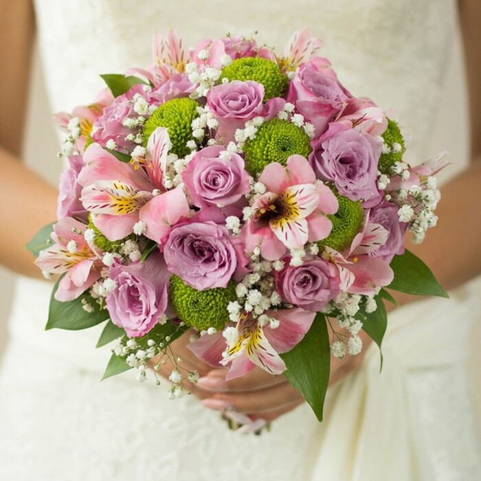 Held gently by a bride whose hands are positioned against the intricate lacework of an ivory gown, this captivating bouquet is a celebration of Tufnell Park's understated elegance. The circular arrangement features silken roses in blush pink and soft lavender, some glistening with tiny beads of moisture, reflecting the freshness of a spring morning after rain on Dartmouth Park's leafy terraces. Vibrant lime green button chrysanthemums interlace throughout the bouquet, their bold hue and rounded form lending energy and textural contrast to the pastel florals. Trumpet-shaped pink alstroemeria, touched with distinctive maroon veining, add intricate detail and an exotic hint, while airy clusters of baby's breath embellish the bouquet with a dreamy softness. Rich, deep green leaves peek from the base, framing the colourful centerpiece in natural sophistication. The bride's simple gold band, visible on one gentle finger, grounds the bouquet in the moment-symbolising new beginnings amid Tufnell Park's community feel. This floral artistry, characteristic of bespoke arrangements found in neighbourhood florists along Fortess Road, encapsulates both romance and contemporary style, making it a perfect choice for modern weddings, bridal inspiration, or as an emblem of meaningful celebration in North London.