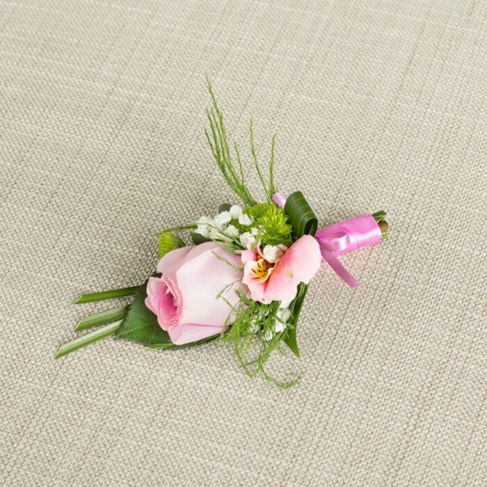 Laid diagonally across a lightly textured, oatmeal-hued linen, this exquisite wedding boutonniere is a true reflection of Tufnell Park's understated elegance. At the heart stands a single, unfurling rose in delicate shades of blush and pale pink, each petal swirling inward to a core tinged with rich, romantic magenta tones. Complementing the rose, a smaller open bloom-its peach petals kissed with yellow and dark feathery markings-mirrors the detail found among vibrant spring gardens near Tufnell Park. Tiny, cloudlike sprigs of baby's breath and plump green chrysanths interlace among an array of foliage: spiky fern fronds, slender blades, and a grounding set of broad, dark green leaves-one deftly folded for drama. A polished, broad leaf elegantly wraps the bundled stems, which are secured with an artful knot of glossy fuchsia ribbon, its bow resting at a jaunty angle and tails trailing gently. Radiant yet soft daylight brings each subtle tone and velvety surface into focus, amplifying the natural beauty of the composition. Intended for a special lapel at a local wedding, this boutonniere suggests sincerity, freshness, and the personable charm of a Tufnell Park ceremony. The combination of refined pastels and lively greens speaks to the neighbourhood's blend of tradition and creativity, offering a singular accent for events both grand and intimate, lovingly prepared by a local florist who honours the community's flair for beauty.