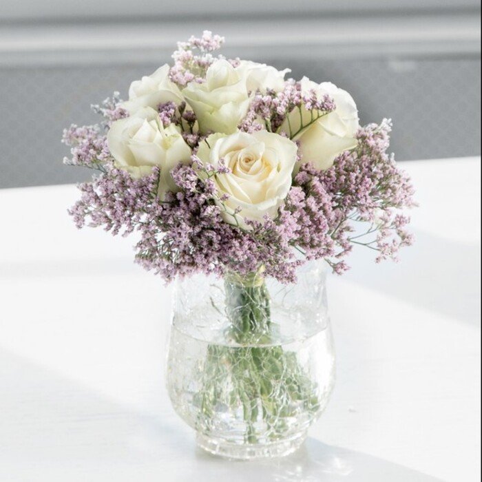 A refined bouquet of creamy white roses and feathery lilac statice occupies the centre of the scene, arranged with the kind of quiet artistry you might expect from a long-established Tufnell Park florist. The flowers form a lush, rounded silhouette, with approximately a dozen white roses as the focal blooms. Each rose displays velvety petals that curl outward in soft layers, the colour hovering between warm ivory and gentle cream, giving an impression of purity without feeling stark. Threaded among the roses are countless tiny blossoms of lavender-toned statice, their papery texture and muted purple hue adding both contrast and volume. These smaller flowers create a misty halo around the larger blooms, filling in pockets of space so that the arrangement appears rich yet breathable, never overcrowded. All of the stems are gathered into a tall, straight-sided glass vase, clear enough to reveal the fresh green stems submerged in clean water, some angled, some almost vertical, hinting at the careful spiral in which the bouquet was hand-tied. The vase sits on a bright white tabletop that reflects the natural light, subtly brightening the underside of the flowers and reinforcing a sense of cleanliness and modernity. The background is blurred into soft, pale tones-suggesting a sunlit room in a Tufnell Park townhouse, perhaps overlooking a leafy street near Campdale Road or close to the quiet stretch towards Dartmouth Park. Light falls gently across the bouquet from a window out of frame, picking up the satin sheen of the rose petals and the more matte, delicate finish of the statice. There is a sense of calm and thoughtful intention here, making this design suitable for many local moments: a peaceful condolence tribute delivered nearby, a simple yet meaningful anniversary gift, or a graceful centrepiece on a sideboard in a North London home that values understated beauty.