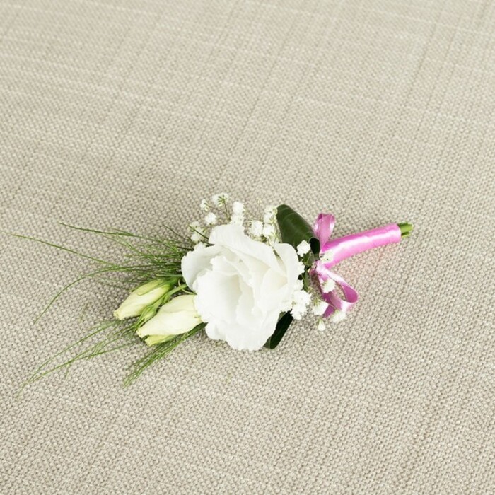 This image shows a meticulously crafted boutonnière resting at an angle across a warm, light beige fabric background that resembles natural linen, its woven texture giving a subtle rustic charm. At the centre of the arrangement is a fully open white lisianthus, also known as eustoma, its petals ruffled and layered into a rounded bloom that feels both delicate and luxurious, like soft silk gathered into gentle waves. Just beneath and slightly to the left of this main flower, two elongated lisianthus buds in creamy green tones sit closely side by side, their tips still closed, adding a graceful sense of growth and continuity, as though symbolising new beginnings for a couple marrying somewhere around Tufnell Park's leafy streets. Sprinkled between the central bloom and buds are airy clusters of tiny white baby's breath, each stem branching into miniature flowers that lend a light, cloud-like fullness to the design. From behind the composition, slender, dark green, grass-like leaves fan out towards the lower left, bringing elegant lines and a touch of movement, while one broader, deeper green leaf rests at the back, grounding the whole piece and making the white petals appear even brighter. The stems are neatly gathered at the lower right and wrapped in a smooth, fuchsia pink satin ribbon, tied into a compact bow that glows vividly against the otherwise muted tones, giving the boutonnière a stylish, modern accent. Soft, even lighting bathes the entire arrangement, highlighting textures without harsh shadows, as if the piece has just been placed on a prep table at a local Tufnell Park studio, ready for delivery to a ceremony near Dartmouth Park. The overall impression is one of understated elegance and fresh romance, ideal for wedding lapels, civil ceremonies, or refined formal events in North London.