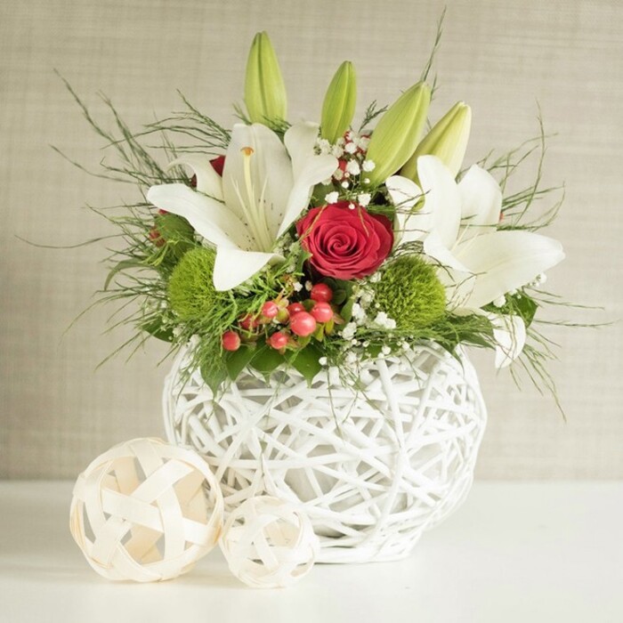 This image captures a luxurious floral centerpiece arranged in a sculptural white woven vase, the kind of stylish design you might see gracing a wedding table in a Tufnell Park restaurant just off Junction Road. The vase is round and hollow, formed from overlapping white strands that create a light, open pattern reminiscent of wicker or artfully twisted branches. It rests on a smooth white surface, its pale colour catching and reflecting the gentle light that falls from the right, while the background fades into a softly mottled beige, allowing the flowers to take centre stage. Emerging in a generous dome from the vase, several pristine white lilies stretch their long stems and unfurl their petals. Some lilies are fully open, their broad, slightly ruffled petals framing golden stamens dusted with pollen, while others remain as elongated green buds, hinting at continued freshness and longevity. Between them, a dramatic red rose sits prominently, its velvety petals layered in a tight spiral, complemented by one or two additional red roses tucked more subtly into the arrangement. The interplay of white and red creates an atmosphere of both serenity and passion, suitable for a wedding breakfast table or an elegant anniversary dinner at home. Bright green, spherical blooms resembling dianthus or chrysanthemums add a fun, pompon-like texture, breaking up the smooth lines of the lilies and roses. Tiny white gypsophila flowers weave delicately through the design like a fine lace, while small red berries introduce further colour and seasonal charm. Cascading greenery, including slender fern-like fronds and broader, darker leaves, frames the arrangement and softens its edges. Two decorative open-weave spheres, crafted from pale natural wood, sit to the left of the vase, mirroring its pattern and enhancing the coordinated, contemporary look. The entire scene radiates thoughtful craftsmanship and quiet celebration, ideal for a refined wedding or intimate gathering in the Tufnell Park area.