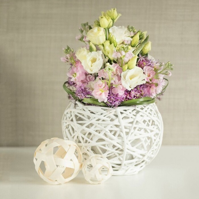 This luminous still life, photographed in a Tufnell Park home where artistry meets comfort, features a floral arrangement that seamlessly blends modern elegance with timeless natural charm. The abundant bouquet, brimming with creamy white and blush yellow lisianthus accented by airy soft pink stock, conjures a pastoral tranquillity. Deep purple filler flowers, likely aster or allium, are nestled at the base, their tiny petals providing an undercurrent of vibrant texture and depth. A border of glossy dark green leaves curves gently around the flowers, highlighting the bouquet's round, lush silhouette. Everything spills from a striking spherical vase, its open-lattice of white painted rattan giving the arrangement a floaty, weightless effect, as if the blooms are suspended mid-air. To the side, two open-weave decorative spheres-fashioned from light wood or natural rattan-sit quietly on a reflective white tabletop, their gentle shadows cast delicately in the bright, indirect light that streams in much like a late morning sun over Tufnell Park's quiet streets. The soft beige backdrop, suggestive of woven wallpaper or a finely textured fabric, adds warmth and tactility, enhancing the arrangement's status as both art and gentle invitation to pause. This composition radiates a sense of renewal-making it ideal inspiration for decorating communal tables at Whittington Park's spring gatherings, or as a centrepiece to elevate family celebrations across north London.