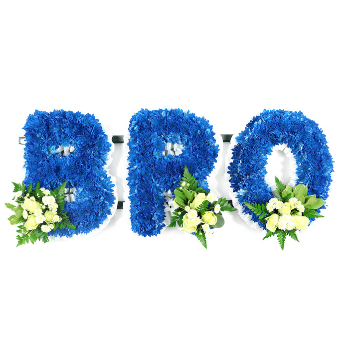 A calm studio image, taken at eye level on a bright white background, shows a three-letter sympathy tribute spelling BRO, each letter formed from a dense mass of vivid royal blue chrysanthemum blooms. The flowers are packed so tightly their rounded heads create a plush, tactile surface, while a slim white foam frame edges each character and defines the inner loops and hollows-a practical structure visible where petals meet negative space. Placed at the lower left of the B and towards the lower right of R and O, delicate clusters of pale ivory or creamy yellow spray roses nestle among small white filler blooms-mini chrysanthemums or baby's breath-lifting the design with gentle highlights. Interspersed greenery, including broader glossy leaves such as salal or lemon leaf and finer fern fronds, threads through the accents to add depth, subtle fragrance, and a cooling green contrast to the saturated blue. The studio's neutral light softens shadows and accentuates the texture of petals, giving the letters a serene, almost tactile presence. Emotionally the piece is composed and respectful: the strong blue suggests fidelity and calm, the cream roses signify consolation, and the visible construction shows careful funeral floristry meant to honour a brother or cherished friend. This arrangement would sit with quiet dignity at a memorial or wake, crafted to be readable and heartfelt from the front pew to the graveside, and to offer a visual language of remembrance that comforts mourners.
