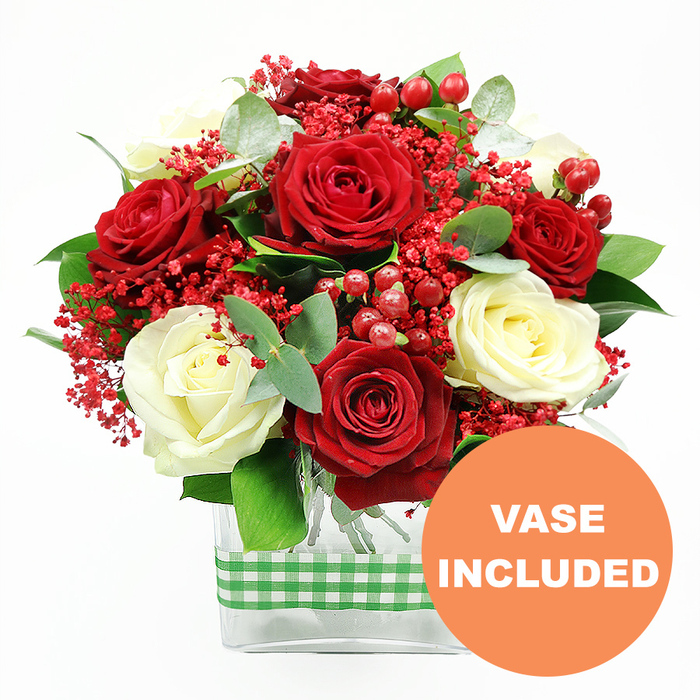 The image presents a refined floral arrangement standing alone against a bright white backdrop, allowing every colour and texture to stand out clearly. Dominating the composition is a lush gathering of red and white roses, their blooms packed closely together to form a generous, almost domed shape. The red roses are rich and velvety, deep in tone, suggestive of classic romance, while the creamy white roses offer a softer, more tranquil note, their gently unfurling petals catching the light with a subtle sheen. Between these focal flowers, tiny clusters of vivid red filler blossoms, reminiscent of dyed gypsophila, drift like a fine mist throughout the arrangement, softening the interplay between the larger heads and adding a cheerful, holiday-like sparkle. Nestled amongst them are rounded red hypericum berries, their polished skins introducing a sculptural quality that contrasts with the softness of the petals. Surrounding and supporting this central mass are sprigs of eucalyptus and other green foliage in different shapes and hues, from broad, deep green leaves to lighter, silvery-green accents that hint at cool, fresh fragrance. All of these elements are carefully set into a clear rectangular glass vase at the base of the image. The stems inside are visible and neatly aligned, reinforcing the sense of professional care and order. Around the middle of the vase, a green and white gingham ribbon wraps horizontally, adding a note of rustic comfort that would feel perfectly at home in a Tufnell Park kitchen overlooking the streets near Junction Road. In the bottom right corner of the picture, an orange circle with white capital letters reads VASE INCLUDED, signalling that this is a complete, ready-to-display bouquet, ideal as a thoughtful gift for anniversaries, Valentine's Day, or simply brightening a living room in North London with a touch of seasonal elegance.