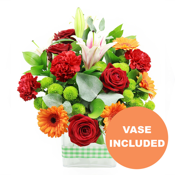 A joyful, richly coloured floral arrangement is displayed in a sleek, clear glass vase, photographed on a pure white background so the bouquet appears almost to glow. The vase is rectangular and contemporary, wrapped neatly at the base with a light green and white gingham ribbon that adds a cheerful, slightly rustic finish, reminiscent of picnics on Hampstead Heath or relaxed weekends in nearby Waterlow Park. Over the rim spills a generous mix of seasonal blooms, carefully balanced in height and colour. Deep crimson red roses, with velvety petals layered tightly around their centres, provide a sense of romance and depth. Alongside them, ruffled red and orange carnations add a lively, frilled texture that softens the strong lines of the design. Bold orange gerbera daisies, each framed by flat, evenly spaced petals and centred with dark chocolate-brown disks, offer a bright, sunny contrast that feels instantly uplifting. Threaded through the arrangement are clusters of small, perfectly round lime-green pompom chrysanthemums, like tiny mossy spheres that bring freshness and visual rhythm. Rising above these lower blooms are white Oriental lilies, some fully open with flared, star-shaped petals traced with subtle pink stripes, others still enclosed in sleek buds poised to unfurl. Their presence gives the arrangement height, elegance, and a gentle sense of movement. Deep green foliage and sprigs of silver-grey eucalyptus loop around the flowers, adding structure and a cool, aromatic note that suggests natural woodland edges beyond Tufnell Park's streets. In the lower right of the image, overlapping the vase, a circular orange badge features the words VASE INCLUDED in bold white capitals, making it clear that this modern glass vase is part of the gift, ready to be delivered and placed straight onto a table, desk, or windowsill in homes and offices across Tufnell Park. 