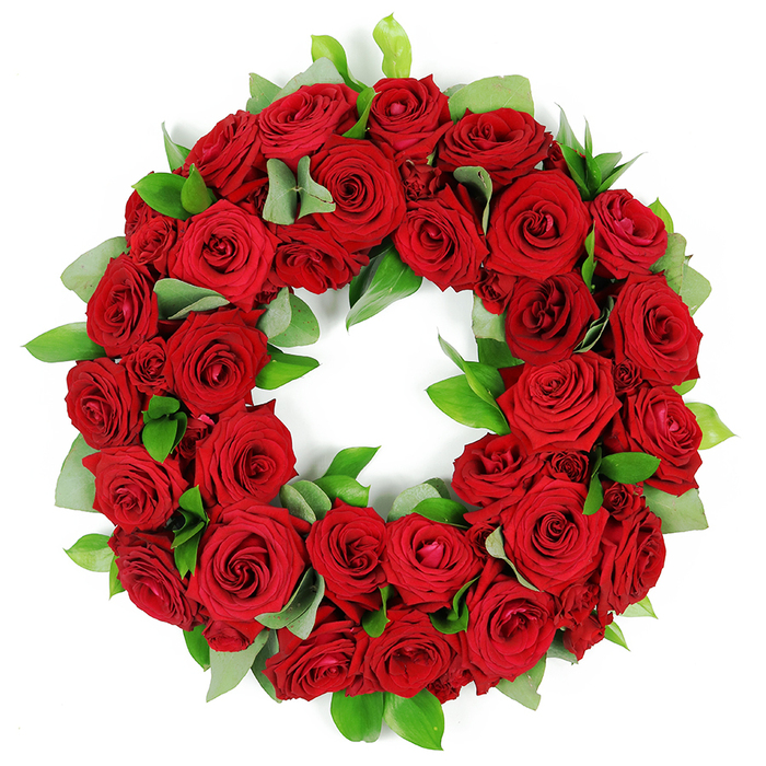 A deeply symbolic circular floral wreath, meticulously crafted from dozens of vibrant, deep red roses, rests against a white background. The blooms are densely packed, forming a lush tapestry of crimson that ranges from closed buds to fully unfurled petals, each petal with rich folds and saturated color. Sprigs of fresh green foliage weave through the roses-broad eucalyptus-like leaves and narrow, glossy ruscus-creating a striking textural contrast that heightens the depth of the arrangement. The wreath projects a three-dimensional quality, with petals and leaves layered to give fullness and presence. This elegant red rose wreath conveys solemn beauty, heartfelt emotion, and enduring tribute, a powerful visual element suitable for memorial services, funeral arrangements, or commemorations. Set in the context of North London, it feels at home in Tufnell Park and the surrounding neighborhoods, a thoughtful expression from a local florist to families marking anniversaries or paying respects. When time allows, same-day delivery is available across the area, bringing a sense of convenience and care to a difficult moment.