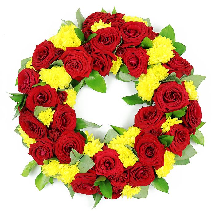 The image shows an elegant, full circular funeral wreath laid out against a crisp white background, so every detail of the flowers and foliage can be clearly seen. The wreath is dominated by luxuriant deep red roses, each one fully open with perfectly formed, velvety petals spiralling inward, creating a sense of depth and quiet intensity. The roses vary subtly in tone, some leaning towards rich crimson, others towards a darker wine red, giving the design a natural, hand-crafted character rather than a flat, uniform look. Interwoven throughout the ring are generous clusters of bright yellow chrysanthemums, likely spray or button varieties, whose compact, feathery petals add a gentle softness and a cheerful glow. These yellow blooms create striking pockets of light between the darker roses, symbolising hope and gratitude amid grief. Surrounding and cradling the flowers, dense green foliage shapes and supports the circle: broad, shiny leaves that could be salal or ruscus sit alongside finer, grey-green sprigs reminiscent of eucalyptus. The leaves fill the spaces without clutter, giving the wreath a lush, rounded profile and a distinctly natural feel, as if it were inspired by the greenery in nearby Tufnell Park gardens and tree-lined streets. The entire arrangement forms a complete, unbroken ring with a clear hollow centre, evoking eternity, continuity and lasting remembrance. Even, shadow-free lighting highlights the freshness of every petal and leaf, mirroring the care an experienced Tufnell Park florist would use when preparing a sympathy tribute for a local funeral service, memorial gathering, or quiet graveside visit.