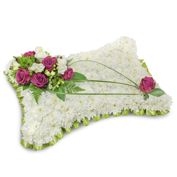 A classic pillow-style funeral tribute photographed on a pristine white field, its surface completely covered with hundreds of small white chrysanthemum florets that together read as a soft, cloud-like mattress of blooms. The chrysanthemums' dense, cushiony texture suggests stillness and repose, while a neat scalloped trim of pale lime-green satin ribbon frames the base with a gentle, restorative hue and a subtle shimmer. Crowning the top-left is an artful cluster of velvety deep magenta roses, their rich petals unfurling like quiet declarations of affection; intermingled are slender white freesia or creamy lisianthus blooms and feathery fern fronds that introduce lightness and a whisper of fragrance. From that focal cluster three long, dark-green bear-grass stems arch gracefully across the white plane, creating a flowing diagonal that leads the eye to a solitary magenta rose placed near the lower-right - a considered echo that completes the visual conversation. The arrangement is lit by soft, even lighting that casts a gentle shadow beneath the pillow, giving it a calm, tangible presence without drama. Prepared with the measured attention Tufnell Park families expect, this sympathy pillow balances formality with tenderness and is often chosen to express condolence and honour at funerals and memorials in local churches and chapels around Dartmouth Park, Fortess Road and the Tufnell Park tube area, reflecting local floral traditions and a quiet, heartfelt craftsmanship.