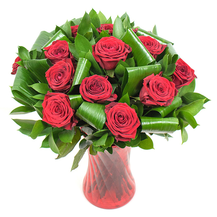 A richly detailed, professionally styled bouquet of deep crimson roses takes centre stage in this image: roughly twelve to fifteen roses, each in full bloom with luxuriously spiralled, velvety petals that deepen to near-burgundy at their hearts. The blooms are densely clustered to form a generous rounded dome, supported and contrasted by a generous mass of dark green foliage. Smaller glossy leaves create a dense, verdant base while larger, bright green leaves-likely Aspidistra or similarly broad foliage-have been intentionally rolled, folded and spiralled into cylindrical and looping shapes. These sculpted leaves produce an architectural counterpoint to the soft rose heads, guiding the eye around the composition and adding modern structure without obscuring the flowers. The bouquet is anchored in a tall, translucent ruby-red glass vase with a subtle vertical rib or twist in the glass, which captures light and adds linear texture that complements the circular forms of the blooms. Placed against a crisp white backdrop, the saturated reds and deep greens stand out with clarity; the even lighting highlights the petals' fine veining and the glossy sheen on the folded leaves. The overall effect is romantic yet contemporary, a statement of refined taste ideal for anniversaries, Valentine's Day, or elevated home décor. Picture this arriving in Tufnell Park after an order placed online from local florists - a tactile gift that balances classic symbolism and modern design, crafted to make an immediate visual and emotional impression in any nearby home or flat overlooking Hillrise or the quieter streets toward Dartmouth Park.