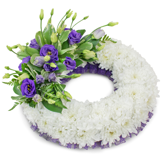 A refined circular memorial wreath set against a bright white background, presented with studio clarity to show material, texture and intent. The wreath's foundation is a lush, continuous ring of white cushion chrysanthemums - innumerable ruffled petals pressed close to one another to form a smooth, cloud-like surface that symbolizes purity and repose. Anchoring the composition in the upper-left quadrant is a lush spray of purple Eustoma (lisianthus), ranging from deep violet, fully open blooms to softer lavender buds that suggest gentle unfolding. The purple cluster is carefully layered with fresh green foliage: glossy, broad leaves with subtle variegation and fine, fern-like greenery that add botanical variety and a sense of natural movement. A subtle trim of dark purple, mesh-like ribbon or lace is visible beneath the chrysanthemums along both inner and outer edges, offering a discreet accent that harmonizes with the lisianthus and lends a composed, finished look. Soft, even lighting and a faint shadow beneath the wreath accentuate the three-dimensionality of the arrangement and reveal the chrysanthemums' fine petal detail. This wreath is intended as a sympathetic tribute for a funeral or memorial, its classic white base and dignified purple accent communicating remembrance, respect, and comfort. When delivered locally - for example to family services or chapels near Junction Road, Fortess Road or the green pockets around Tufnell Park - it presents as a carefully balanced expression of condolence, both traditional and quietly modern in its restrained simplicity.