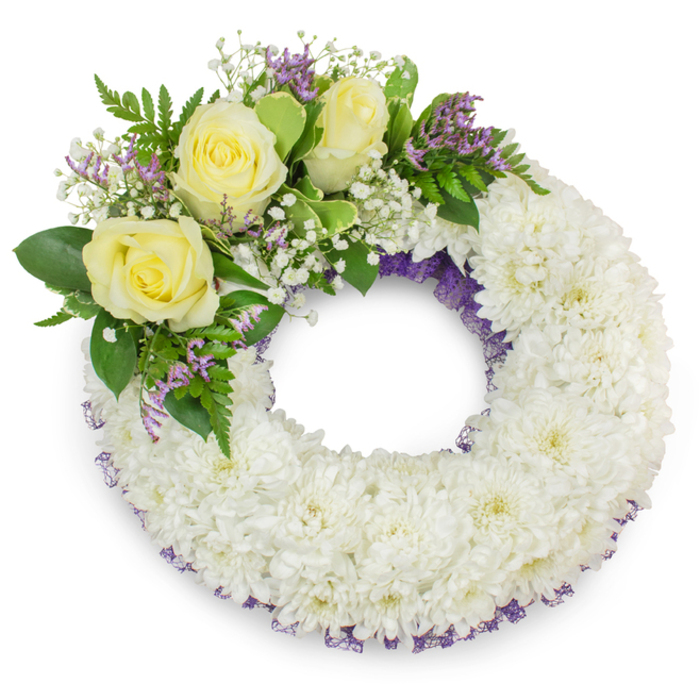 This circular sympathy wreath is presented as a calm, elegant statement of remembrance, photographed against a plain white ground so every detail reads clearly. The foundation is a generous, tightly packed mass of small white spray chrysanthemums; their layered petals create a tactile, cloud-like surface that feels both substantial and gentle to the touch. Along the inner and outer edges a subtle band of deep lavender-purple mesh or ribbon peeks through the white, adding a refined, almost low-key border that gives the wreath depth without overpowering the purity of the chrysanthemums. Set on the wreath's upper-left curve is an intentional cluster of accents: three creamy yellow roses, each opening into soft concentric layers of velvety petals, surrounded by airy sprays of white baby's breath that lend an ethereal, lacy texture. Touches of small, pale purple statice or limonium appear among the foliage, offering a counterpoint of cool colour. The greenery-broad, glossy leaves mixed with finer, fern-like fronds-frames the blooms and brings a living vibrancy to the memorial design. The clear center maintains the traditional symbolism of continuity and unbroken love. The soft, even lighting produces a delicate shadow beneath the wreath, enhancing its three-dimensional form on the white backdrop. This piece, crafted with sensitivity and classical restraint, is the type of tribute often requested by neighbours in Tufnell Park and Kentish Town for chapel services or graveside farewells; Flowers Tufnell Park prepares such arrangements with care, mindful of proportion, scent and the comforting textures that families value.